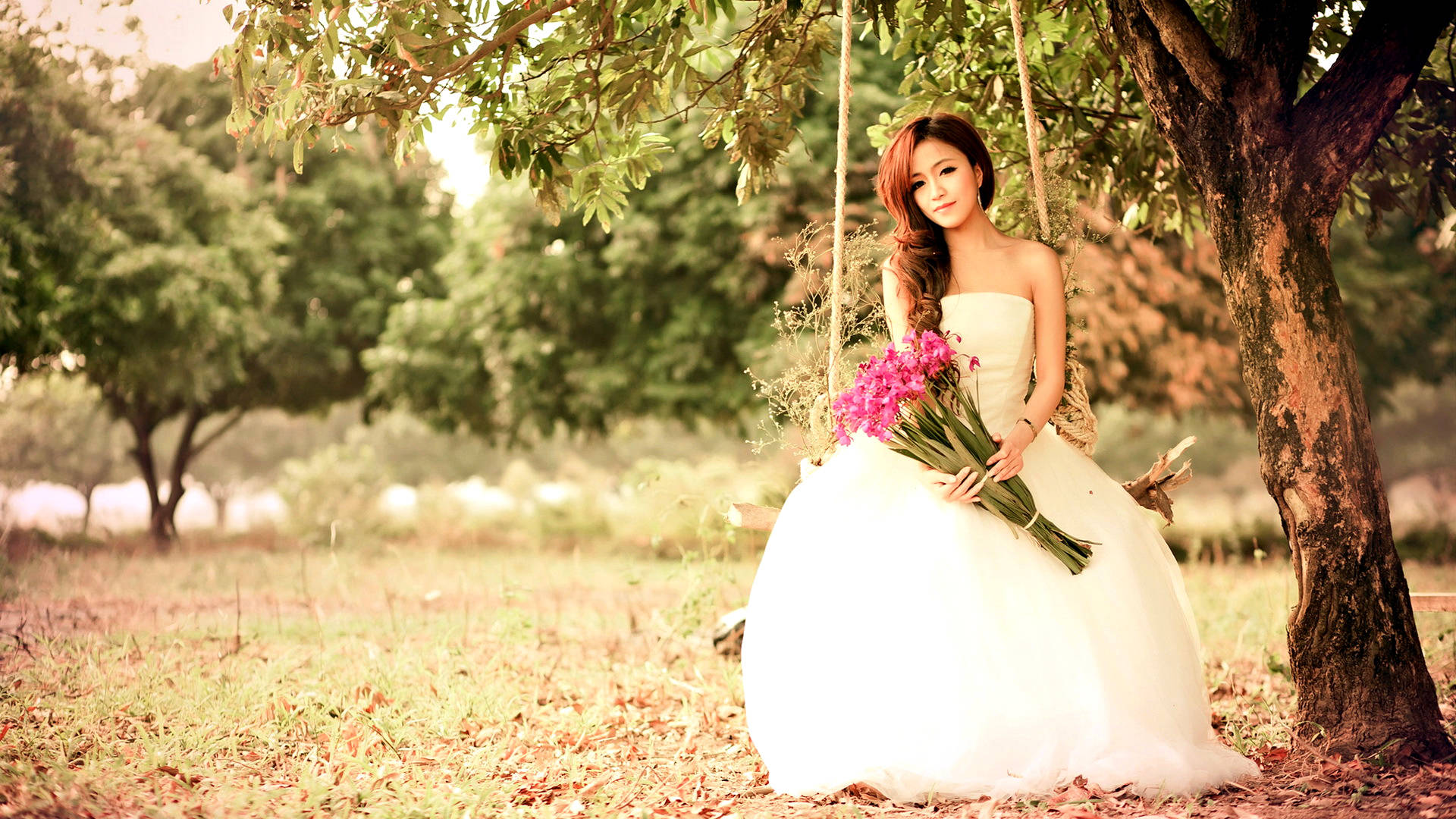 Bride 桌布 壁紙 — 一位身穿白色婚紗的新娘，在寧靜的公園裡，樹影斑駁的陽光下，優雅地坐在樹鞦韆上，手持粉紅色的花朵。