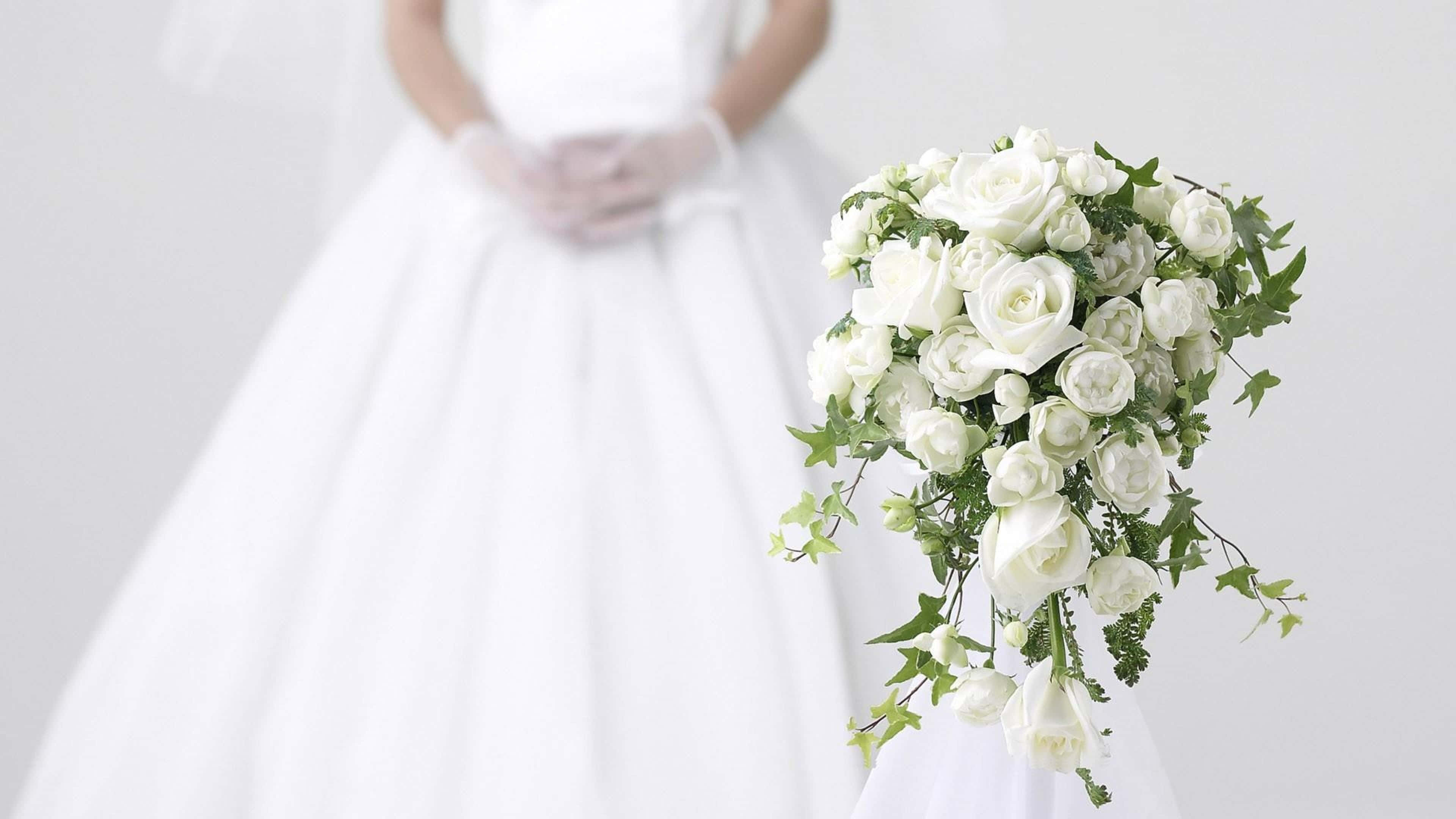 Wedding Bouquet 桌布 壁紙 — 一位身穿白色婚紗的新娘，在柔和的白色背景前，手持一束由白玫瑰和綠葉組成的瀑布式花束。