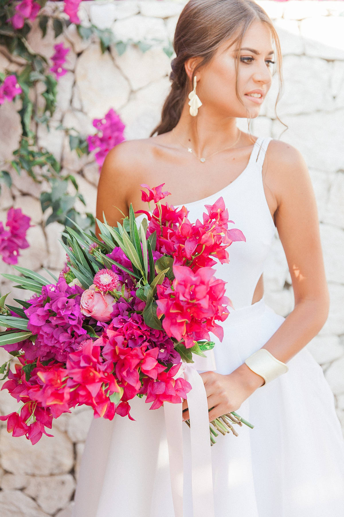 Bougainvillea 桌布 壁紙 — 一位身穿白色婚紗的新娘，在陽光照射的石牆前，手持一束充滿活力的粉紅色和洋紅色花束。