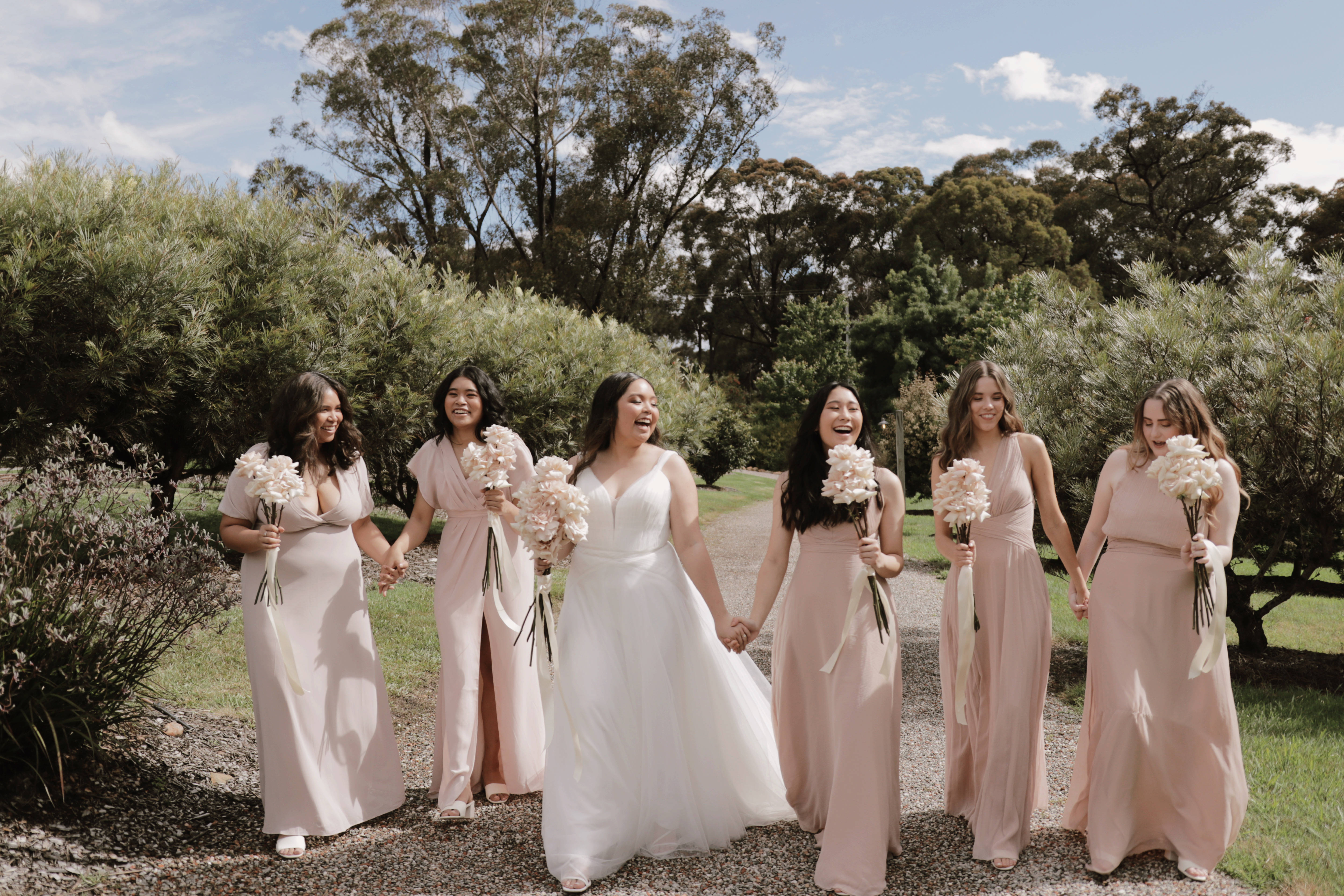 Bridesmaids 桌布 壁紙 — 一位身穿白色婚紗的新娘和她的伴娘們穿著淡粉紅色禮服，在陽光普照的碎石路上手持白色花束，歡快地笑著。
