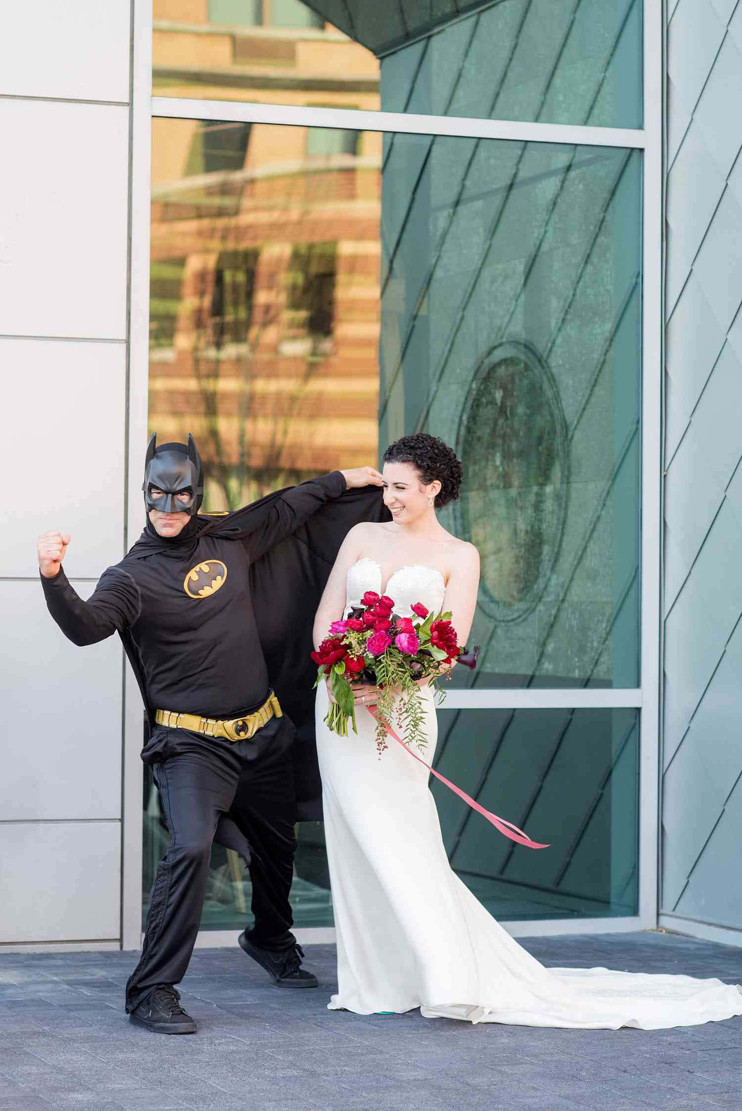 Funny Wedding 桌布 壁紙 — 一位身穿白色婚紗的新娘微笑著，而一名穿著蝙蝠俠服裝的男子在她旁邊，在一棟現代玻璃建築前俏皮地擺姿勢。