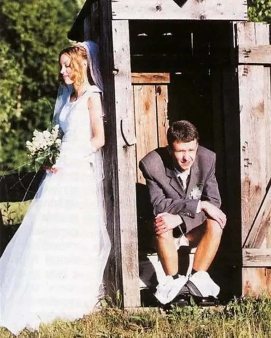 Funny Wedding 桌布 壁紙 — 一位身穿白色婚紗的新娘站在一個質樸的木製戶外廁所旁邊，而她的新郎穿著短褲和襪子坐在裏面。
