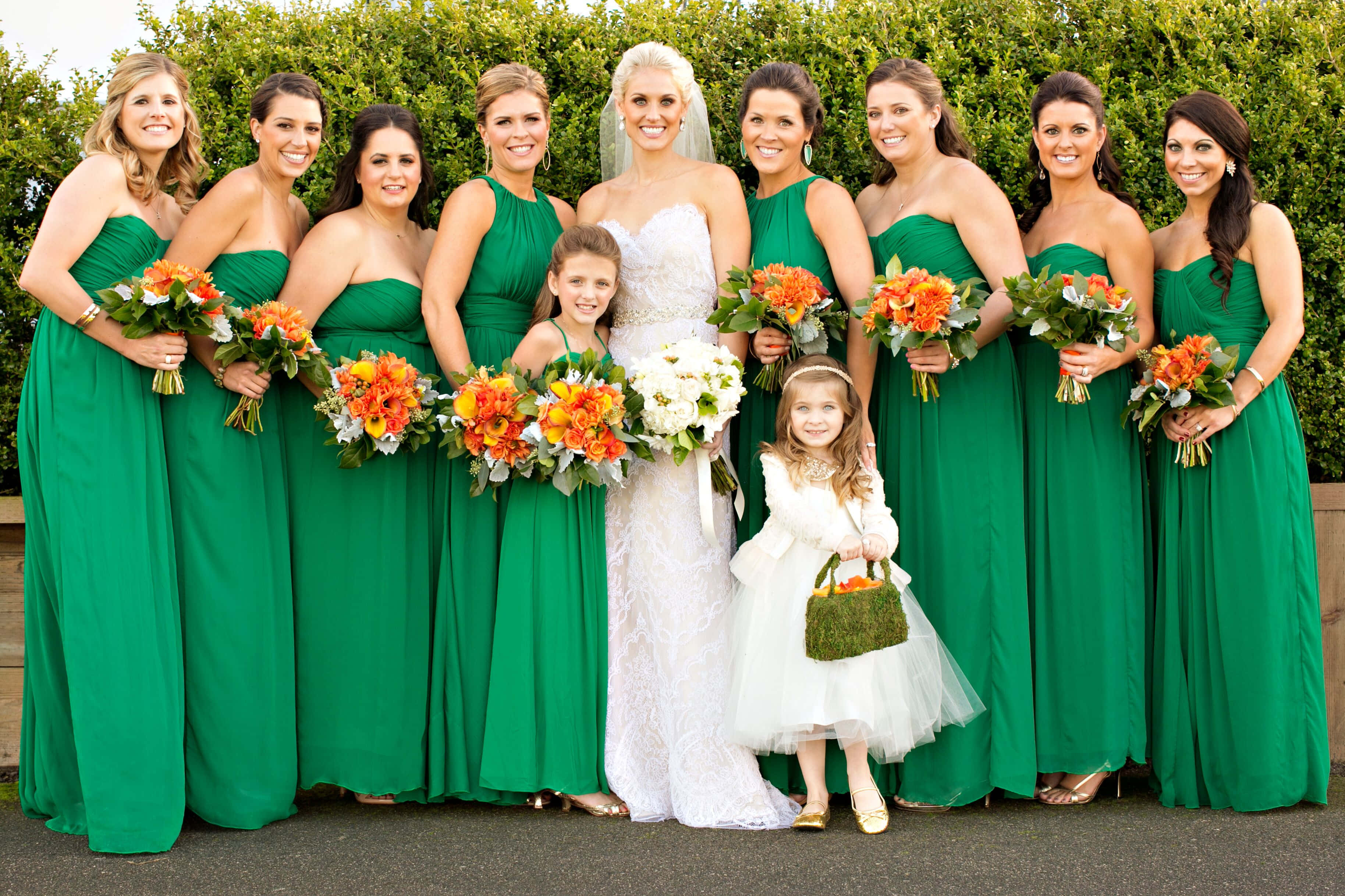 Bridesmaids 桌布 壁紙 — 一位身穿白色婚紗的新娘與微笑的伴娘們穿著翡翠綠色連衣裙，手持橙色花束，站在茂密的綠色樹籬前。