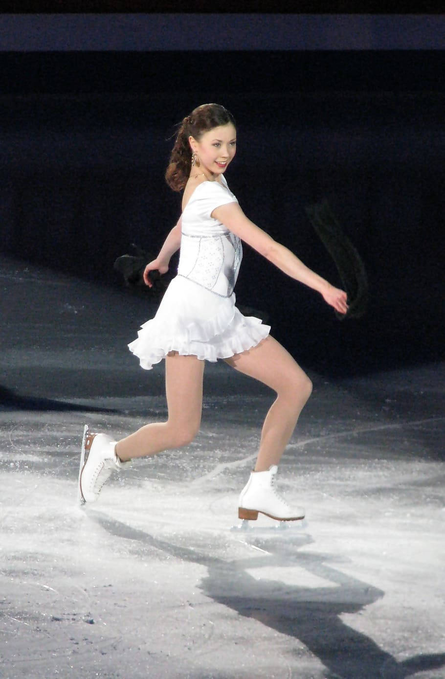 Ice Skating 桌布 壁紙 — 一位身穿白色服裝的花式滑冰女選手在明亮的場館燈光下優雅地滑過冰場。