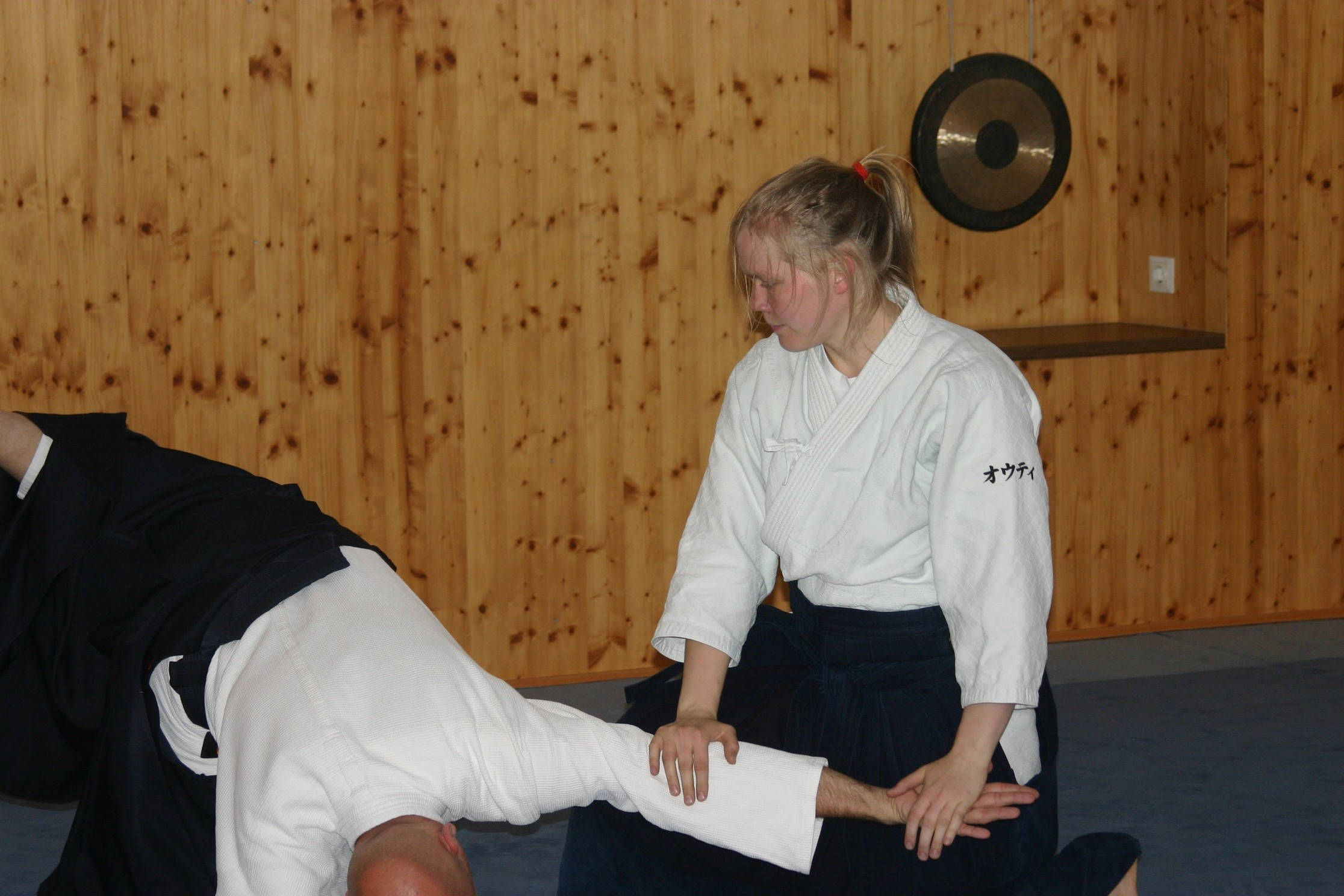 martial arts 桌布 壁紙 — 一位身穿白色柔道服的女性，在一個掛著鑼的木製道場中，對夥伴施展精準的合氣道技術。