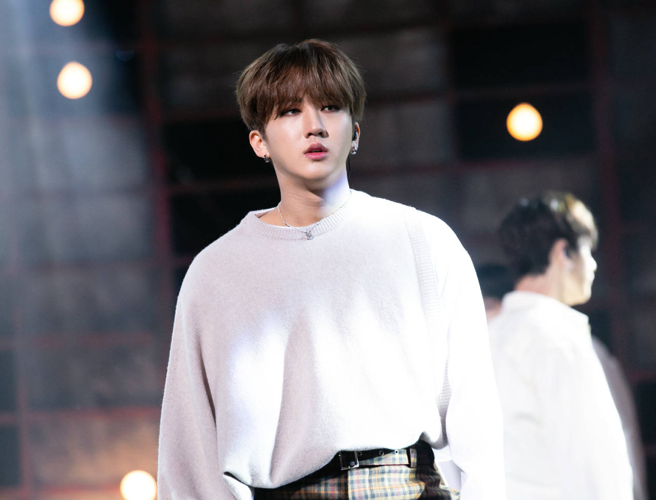 Changbin 桌布 壁紙 — 一位身穿白色毛衣和格子褲的年輕男子自信地站在燈光昏暗的舞台上，身後溫暖的聚光燈閃耀著。