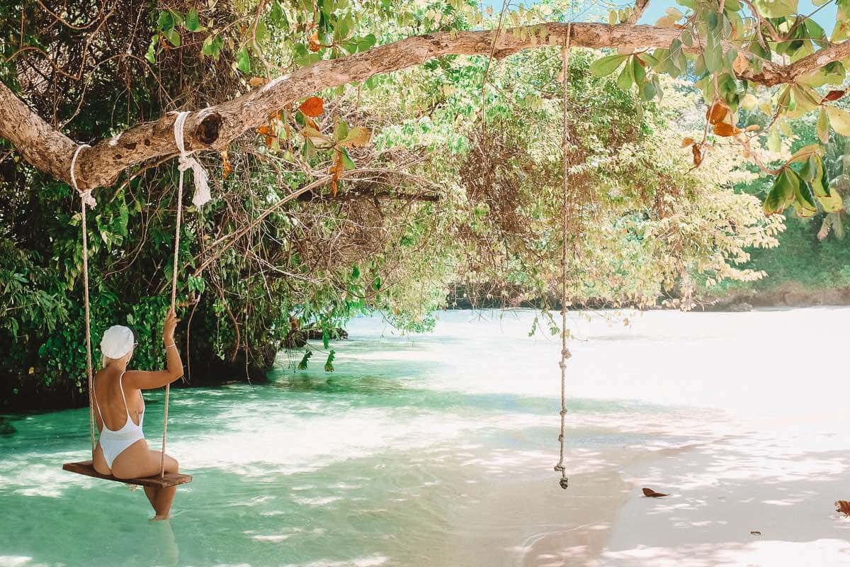 Jamaican Beach 桌布 壁紙 — 一位身穿白色泳衣的女性坐在一個木製鞦韆上，鞦韆掛在熱帶海灘上茂密的樹上，下方是碧綠色的海水。