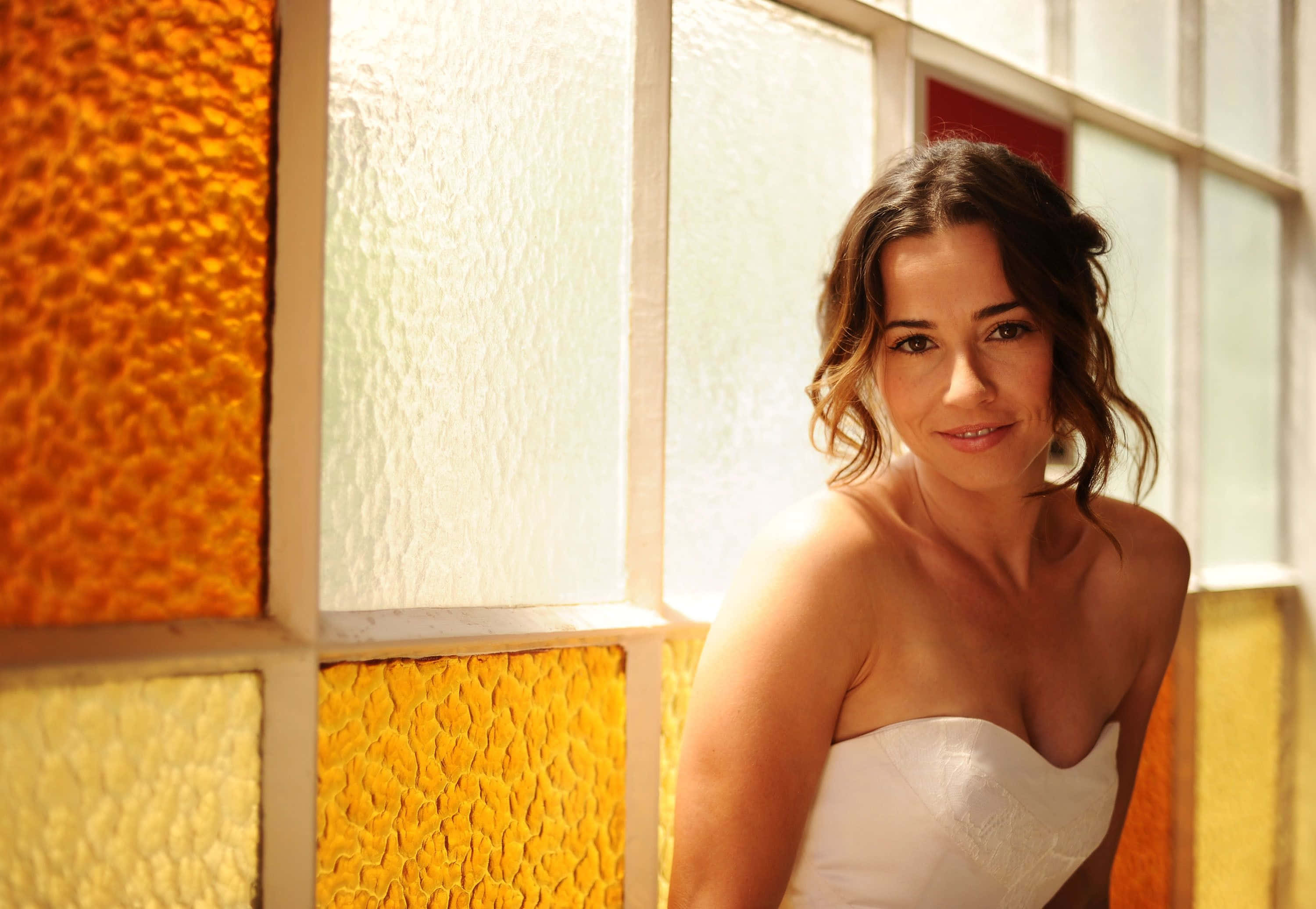 linda cardellini 桌布 壁紙 — 一位身穿白色無肩帶連衣裙的女性在色彩繽紛的紋理玻璃窗旁溫柔微笑。