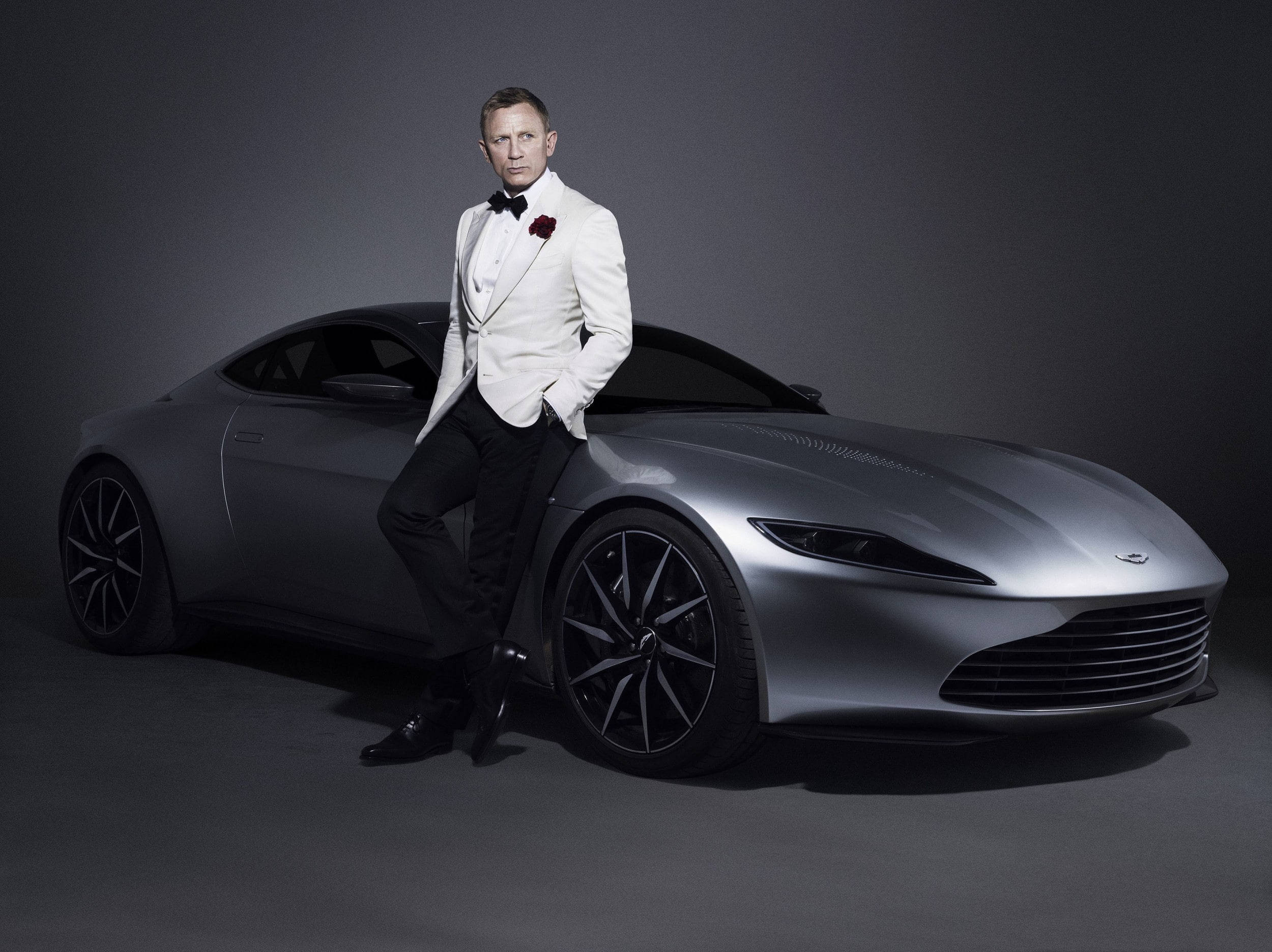 Daniel Craig 桌布 壁紙 — 一位身穿白色燕尾服的男士，在簡約的攝影棚環境中自信地靠在一輛時尚的銀色跑車上。