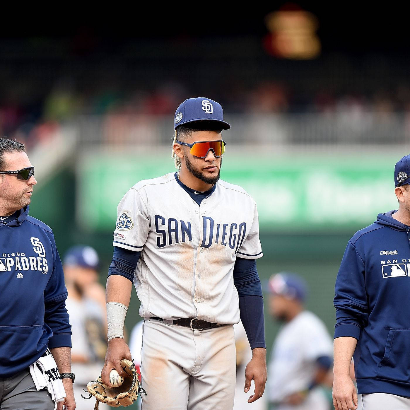 Tatis 桌布 壁紙 — 一位身穿白色球衣、戴著橙色太陽眼鏡的聖地牙哥教士隊棒球選手，在陽光明媚的球場上自信地行走，隊友在附近。