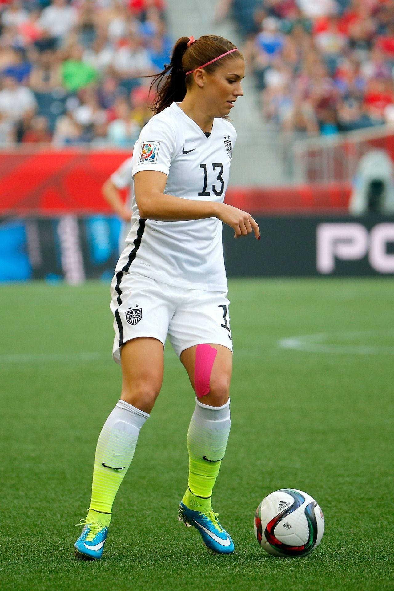 Alex Morgan 桌布 壁紙 — 一位身穿白色球衣、號碼13的女足球員在日間比賽中，於充滿活力的綠色球場上專注地看著足球。