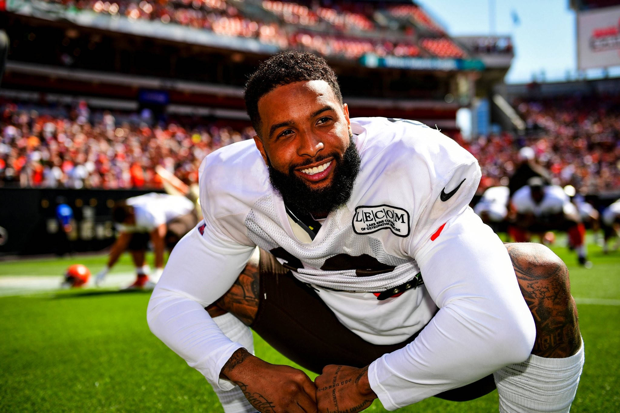 Odell 桌布 壁紙 — 一位身穿白色球衣、面帶微笑的足球員在陽光明媚的球場上蹲下，身後是座無虛席的體育場。