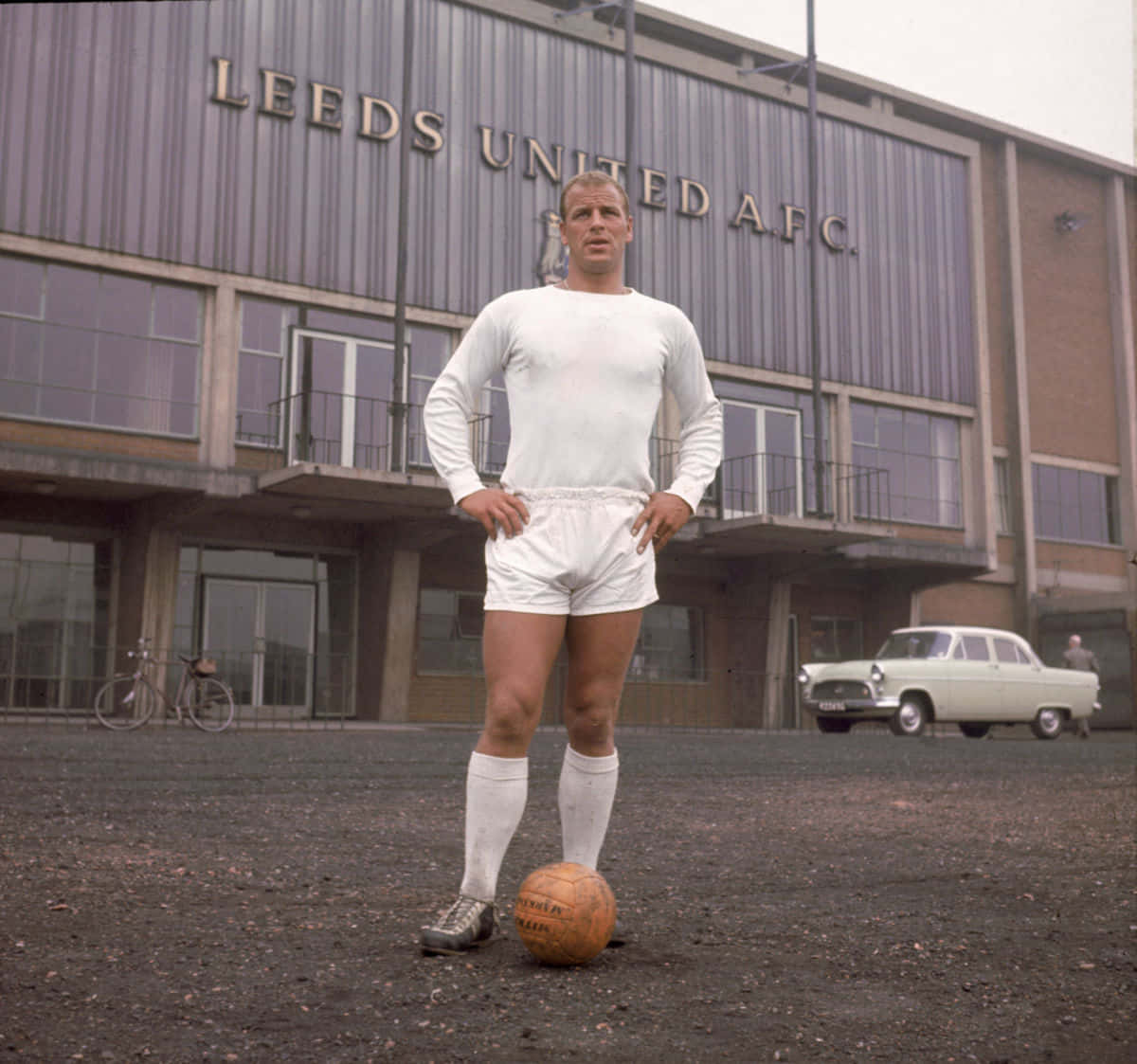 Leeds United Fc 桌布 壁紙 — 一位身穿白色球衣的復古足球員自信地雙手叉腰，站在利茲聯AFC球場前的皮革足球旁。
