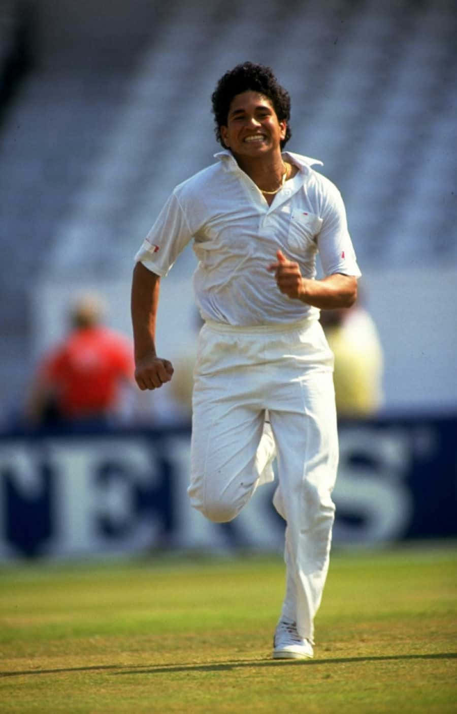 Sachin Tendulkar 桌布 壁紙 — 一位身穿白色球衣的快樂板球運動員在陽光明媚的綠色球場上奔跑，背景是模糊的體育場座位。