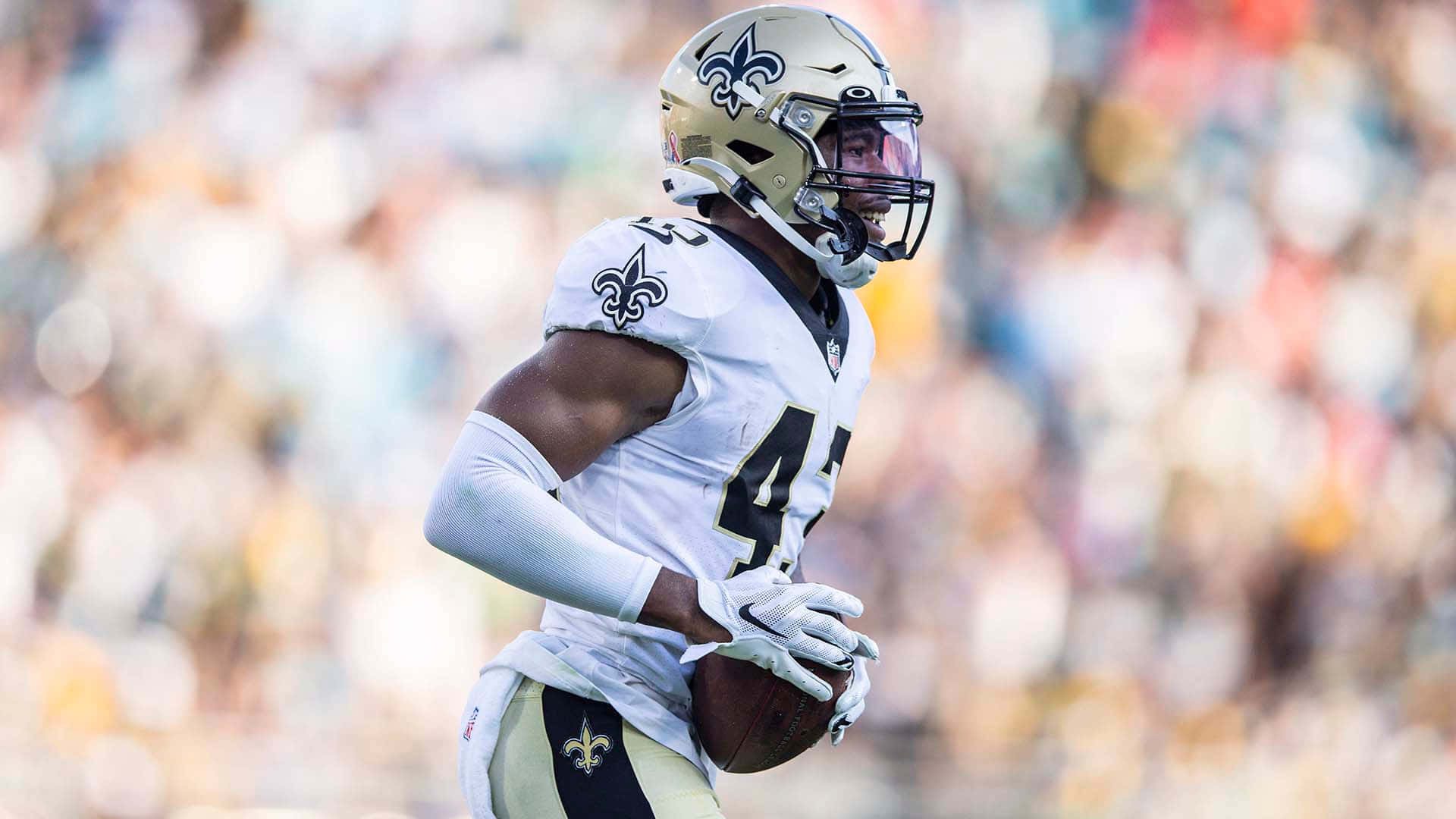 Marcus Williams 桌布 壁紙 — 一位身穿白色球衣的紐奧良聖徒隊美式足球員在日間比賽中帶球奔跑，背景是模糊的體育場人群。
