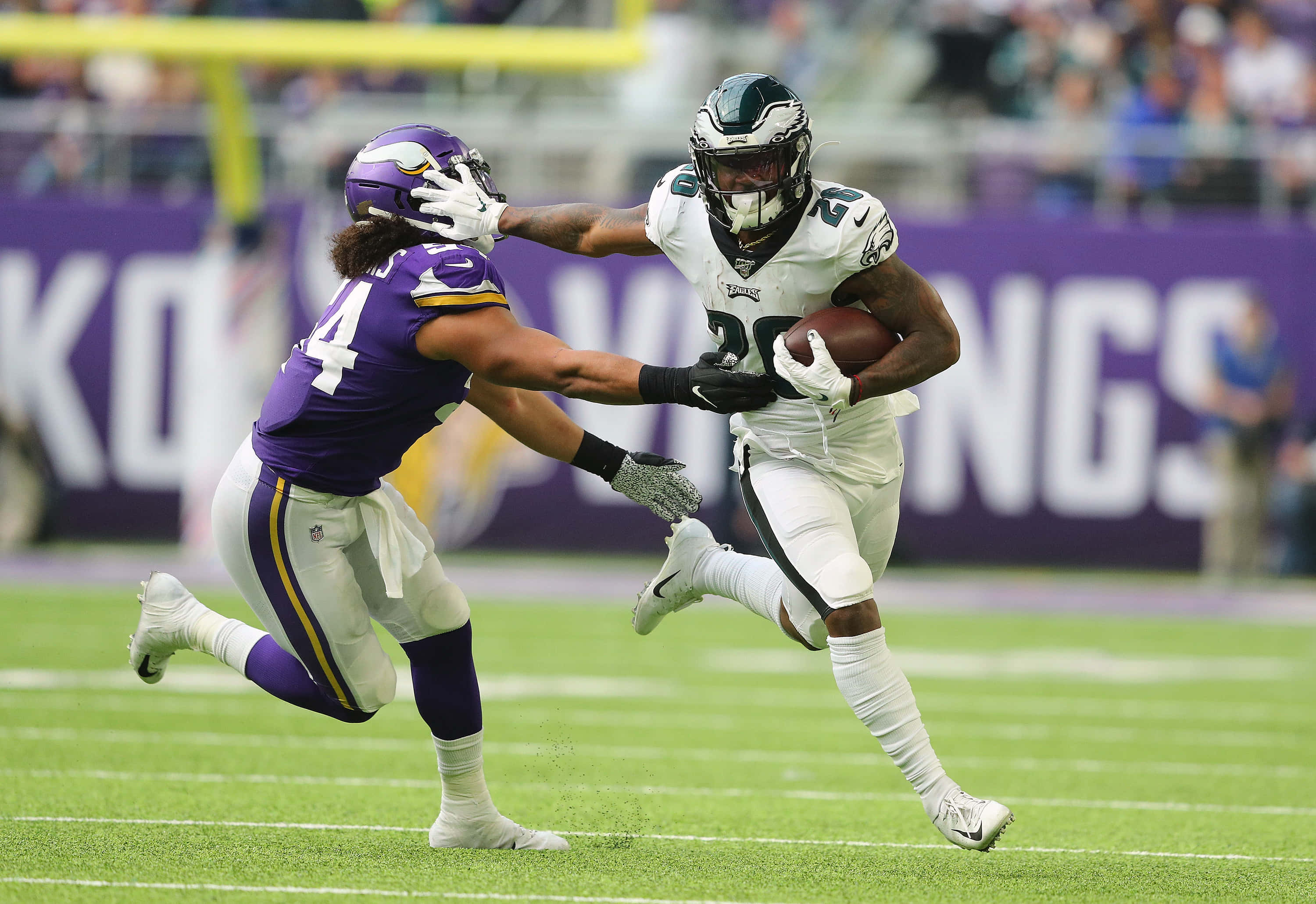 Miles Sanders 桌布 壁紙 — 一位身穿白色球衣的費城老鷹隊跑衛帶著橄欖球衝刺，而一位身穿紫色的明尼蘇達維京人隊防守球員在充滿活力的綠色球場上將他擒抱。