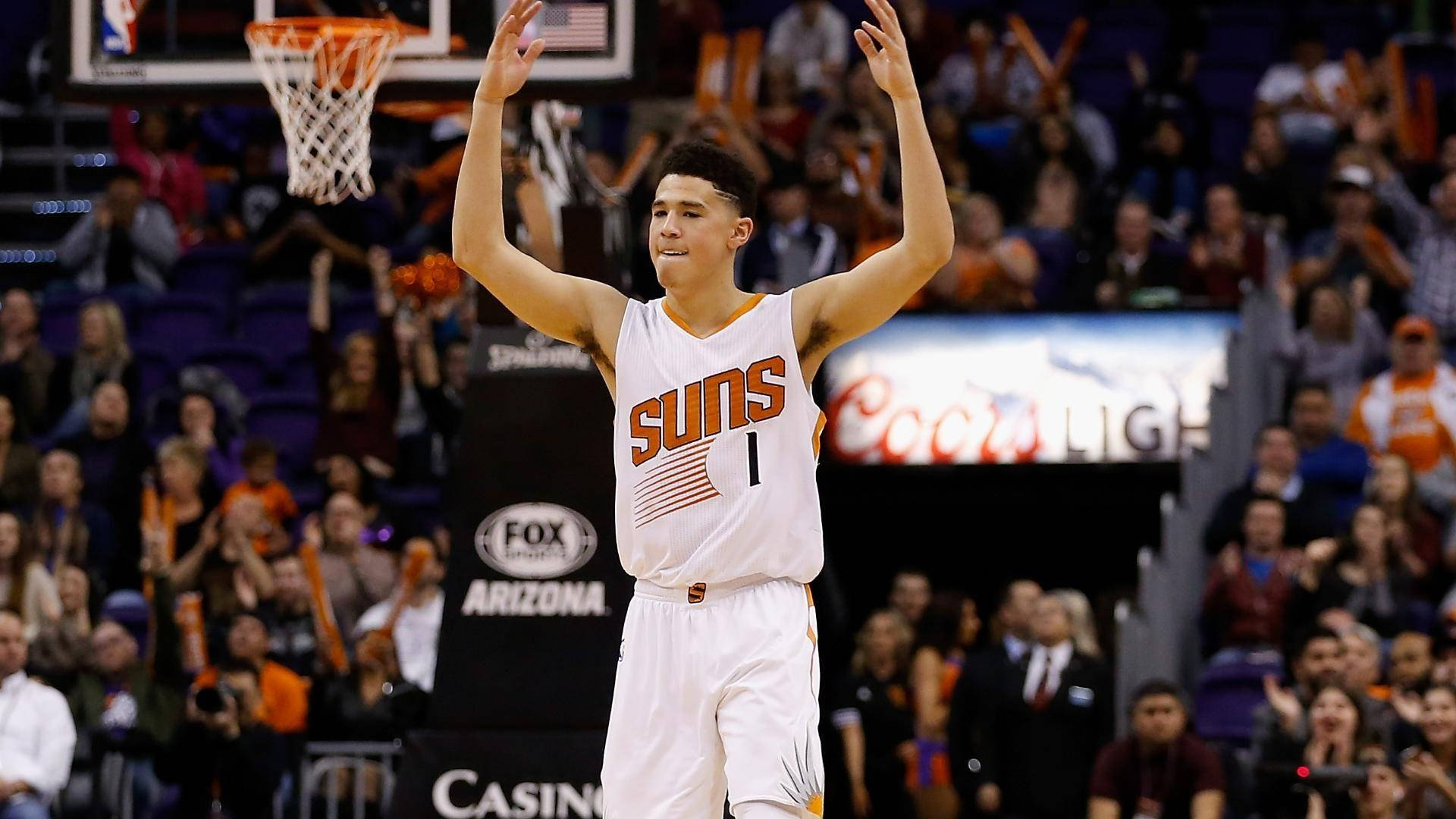 Devin Booker 桌布 壁紙 — 一位身穿白色球衣的鳳凰城太陽隊籃球運動員在明亮的場館燈光下高舉雙臂慶祝，背景中球迷歡呼。