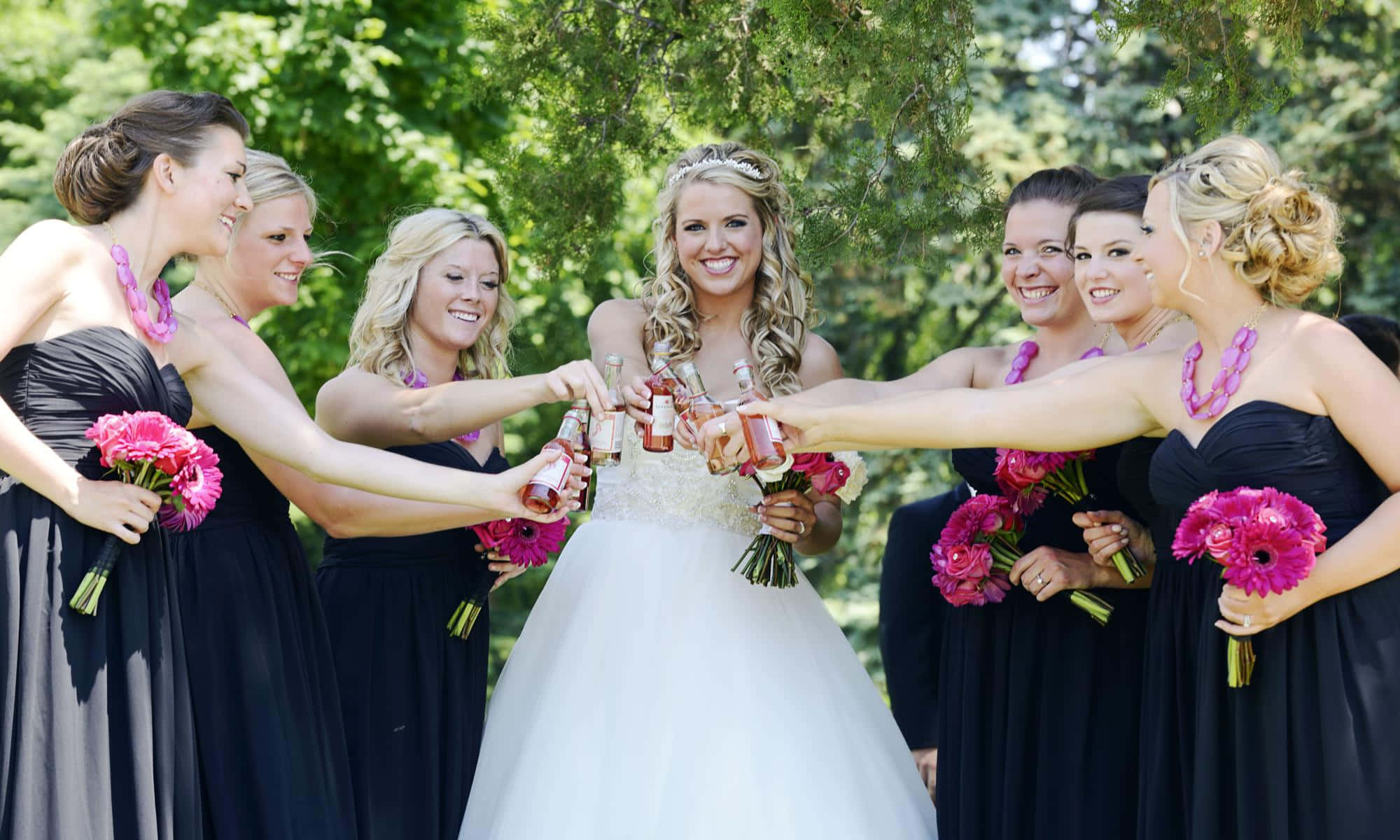 Bridesmaids 桌布 壁紙 — 一位身穿白色禮服的新娘和她的伴娘們身穿黑色禮服，在綠樹成蔭下用玫瑰酒瓶乾杯。