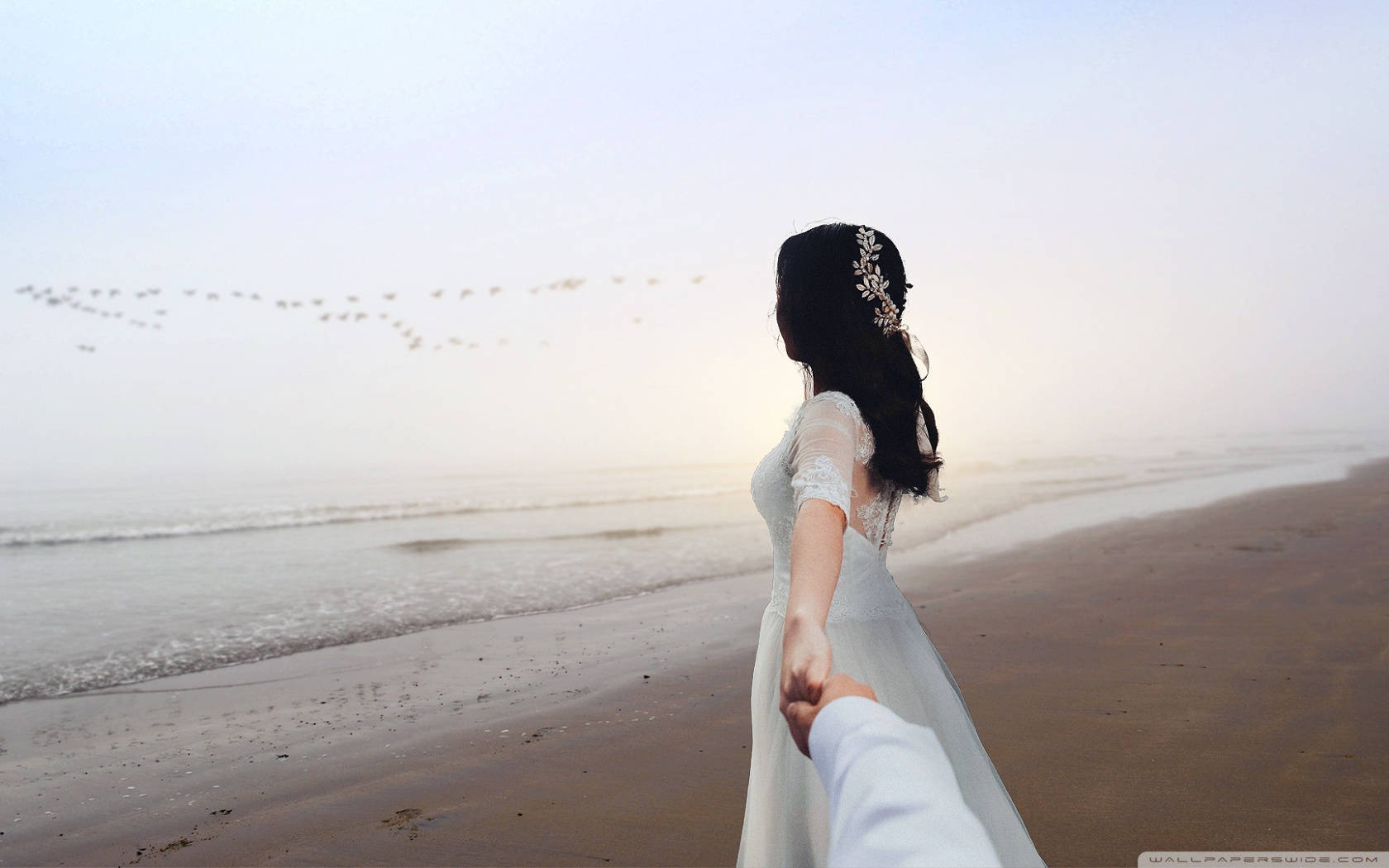Bride 桌布 壁紙 — 一位身穿白色禮服的新娘在日出時分，手牽手沿著霧濛濛的海灘漫步，鳥兒在頭頂飛翔。
