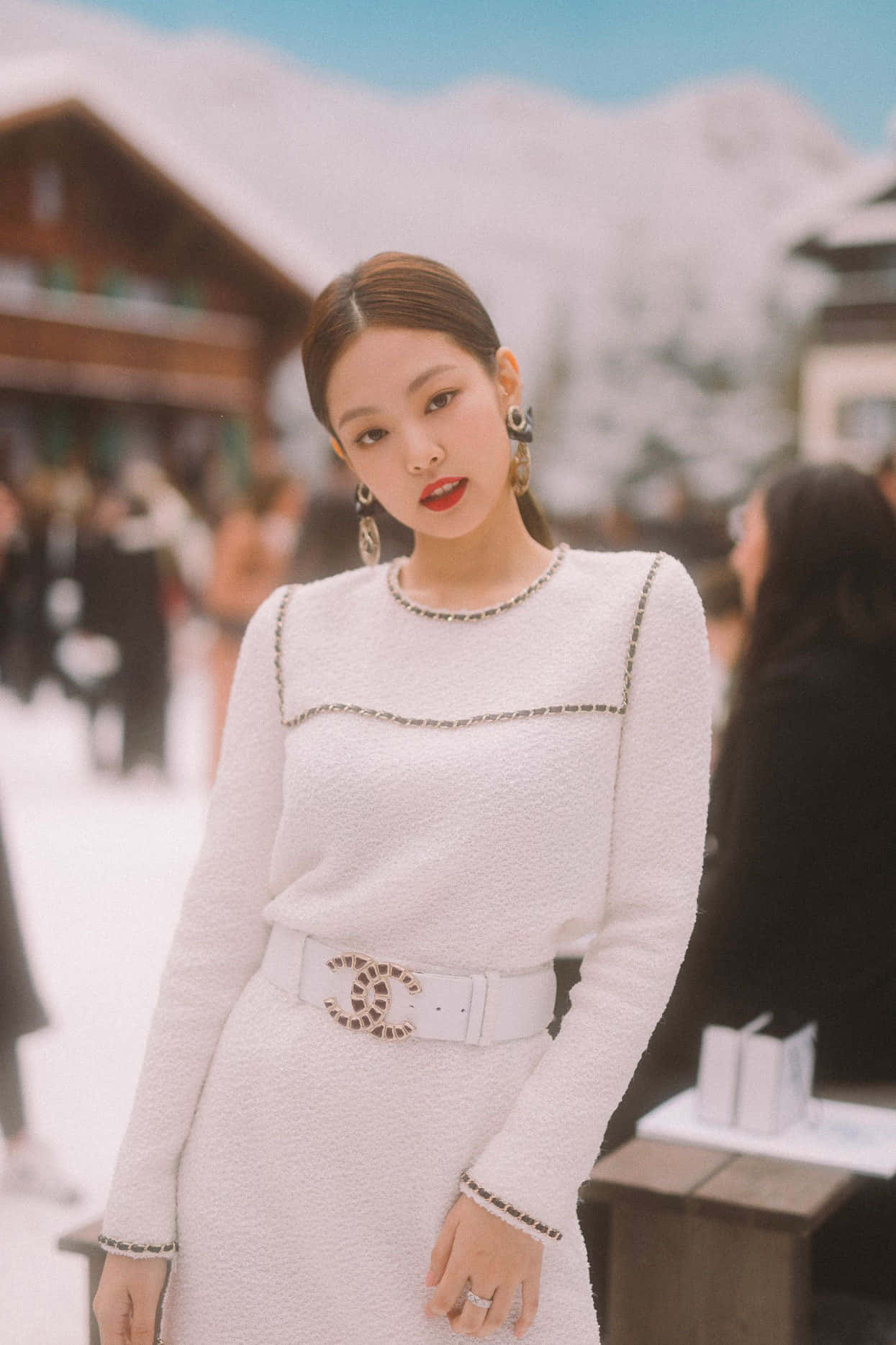 BLACKPINK 桌布 壁紙 — 一位身穿白色粗花呢 Chanel 連衣裙、搭配標誌腰帶的女性，自信地站在白雪覆蓋的山村環境中。