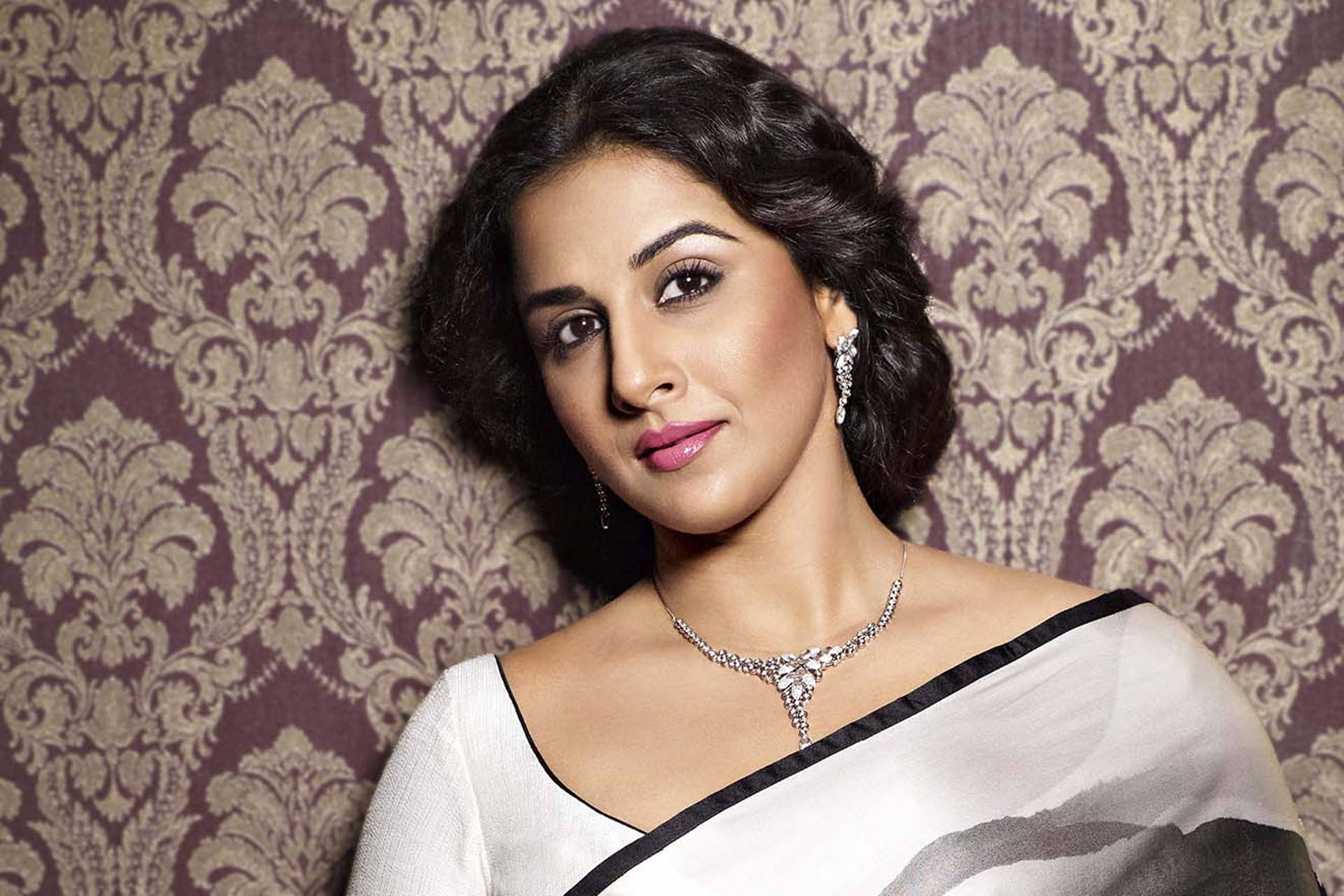 Vidya Balan 桌布 壁紙 — 一位身穿白色紗麗服、帶黑色飾邊和鑽石首飾的女性，在紫色錦緞壁紙前擺姿勢。