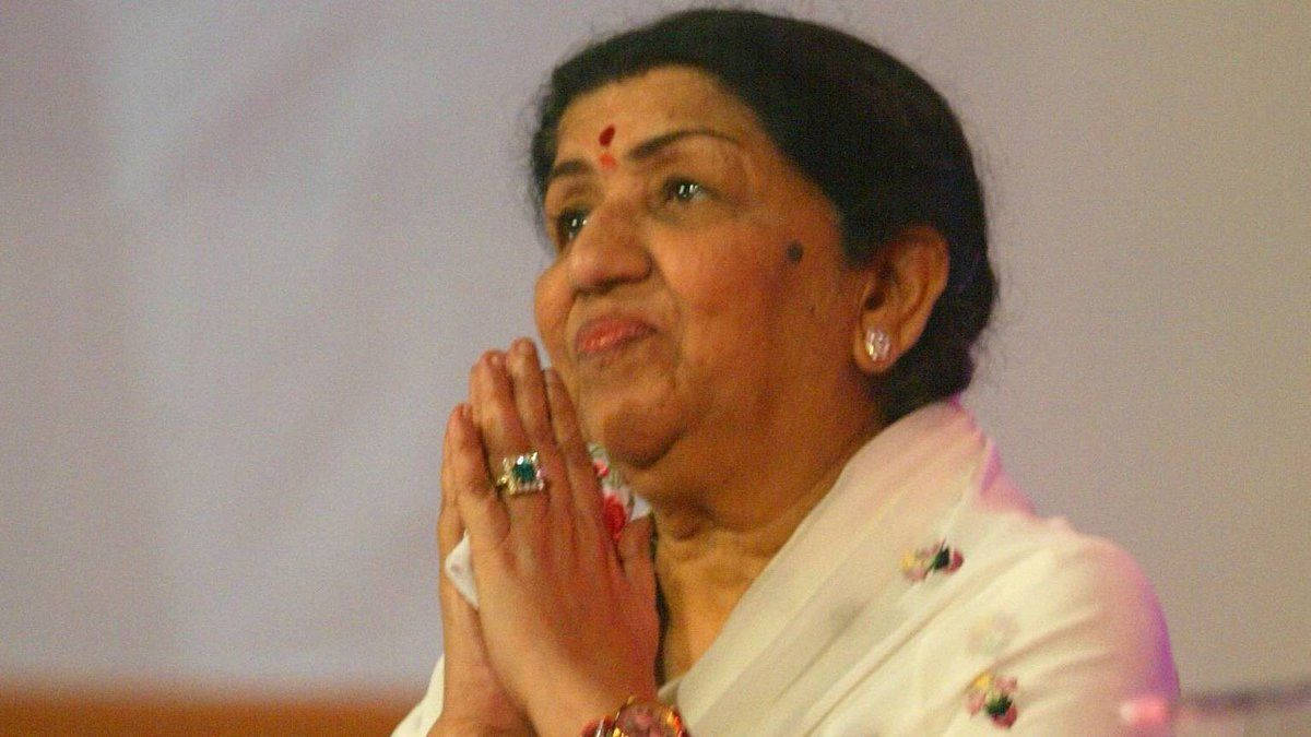 Lata Mangeshkar 桌布 壁紙 — 一位身穿白色紗麗的老年女性雙手合十祈禱，佩戴珠寶和紅色眉心貼，目光向上，表情寧靜。