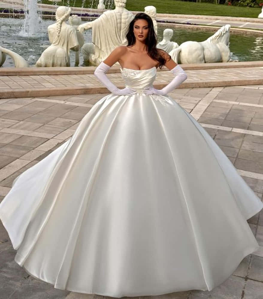 Wedding Gown 桌布 壁紙 — 一位身穿白色緞面大擺舞會禮服的女性，雙手叉腰，自信地站在一座裝飾著古典大理石雕像的噴泉前。