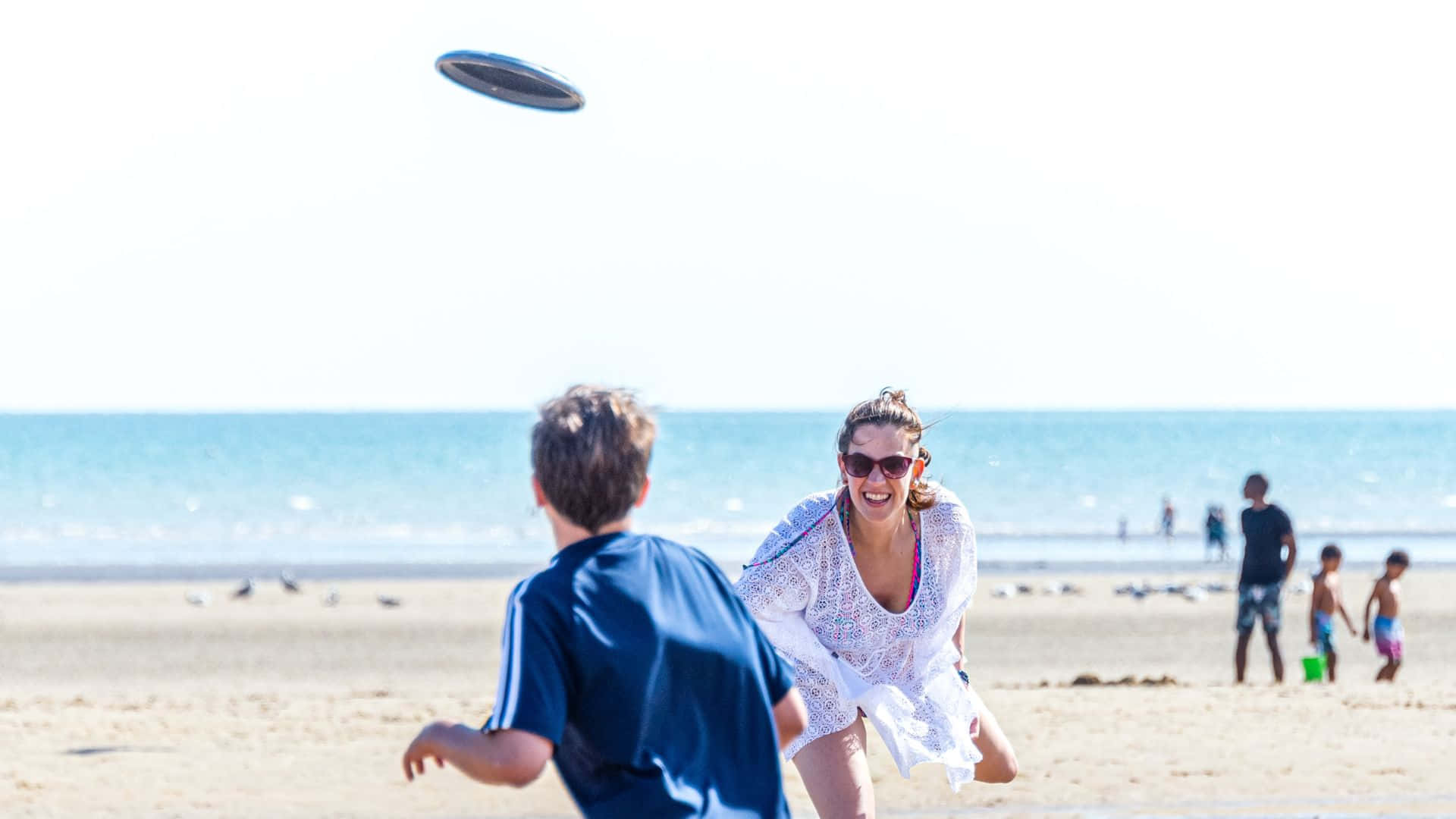 HD Frisbee 桌布 壁紙 — 一位身穿白色罩衫的快樂女性，在陽光明媚的海灘上，身後是碧綠色的海洋，她笑著撲向飛來的飛盤。