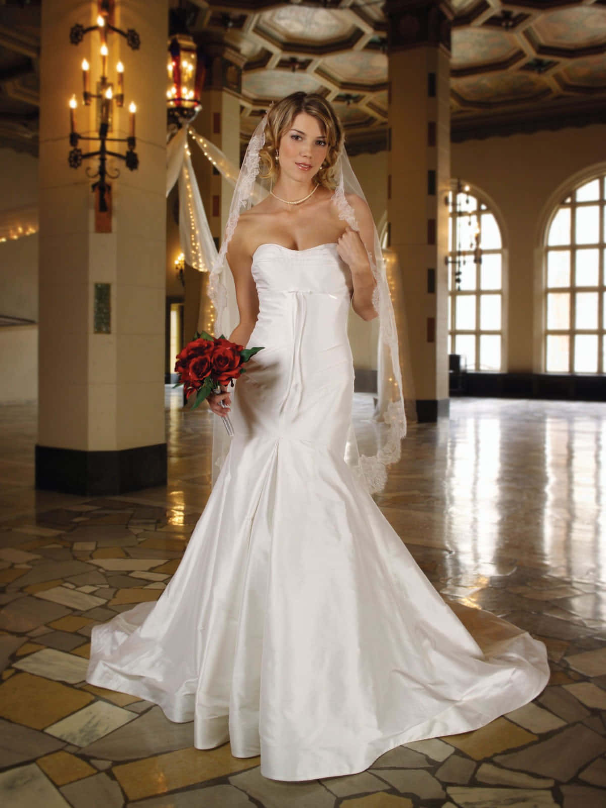 Wedding Gown 桌布 壁紙 — 一位身穿白色美人魚婚紗的新娘在一個有石地板和拱形窗戶的大廳裡拿著紅玫瑰。