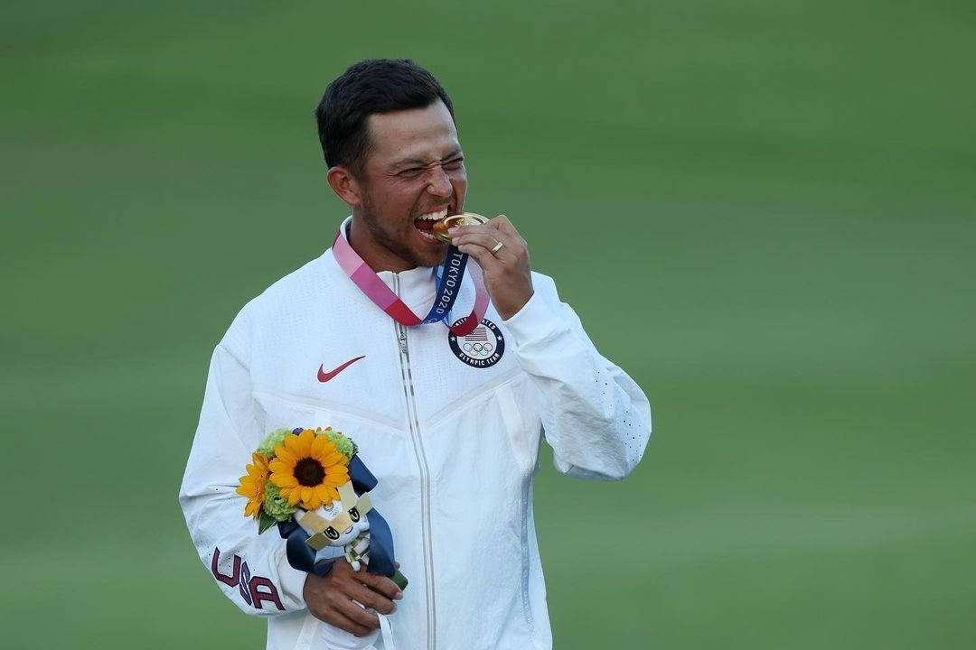 高爾夫 桌布 壁紙 — 一位身穿白色美國外套的奧運選手咬著他的金牌，同時手持向日葵和吉祥物，背景是模糊的綠色場地。