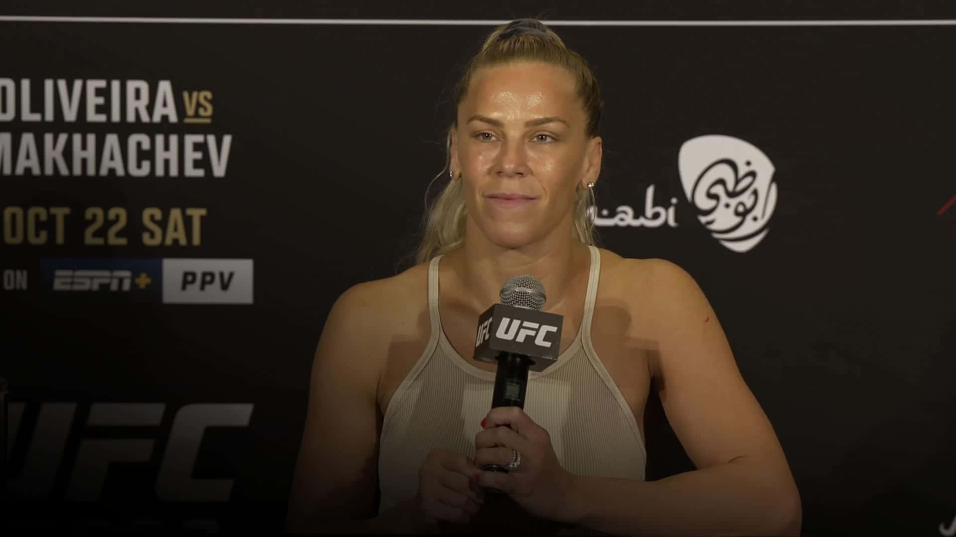 桌布 壁紙 — 一位身穿白色背心的女性UFC拳手在'Oliveira vs Makhachev'背景的新聞發布會上拿著麥克風。