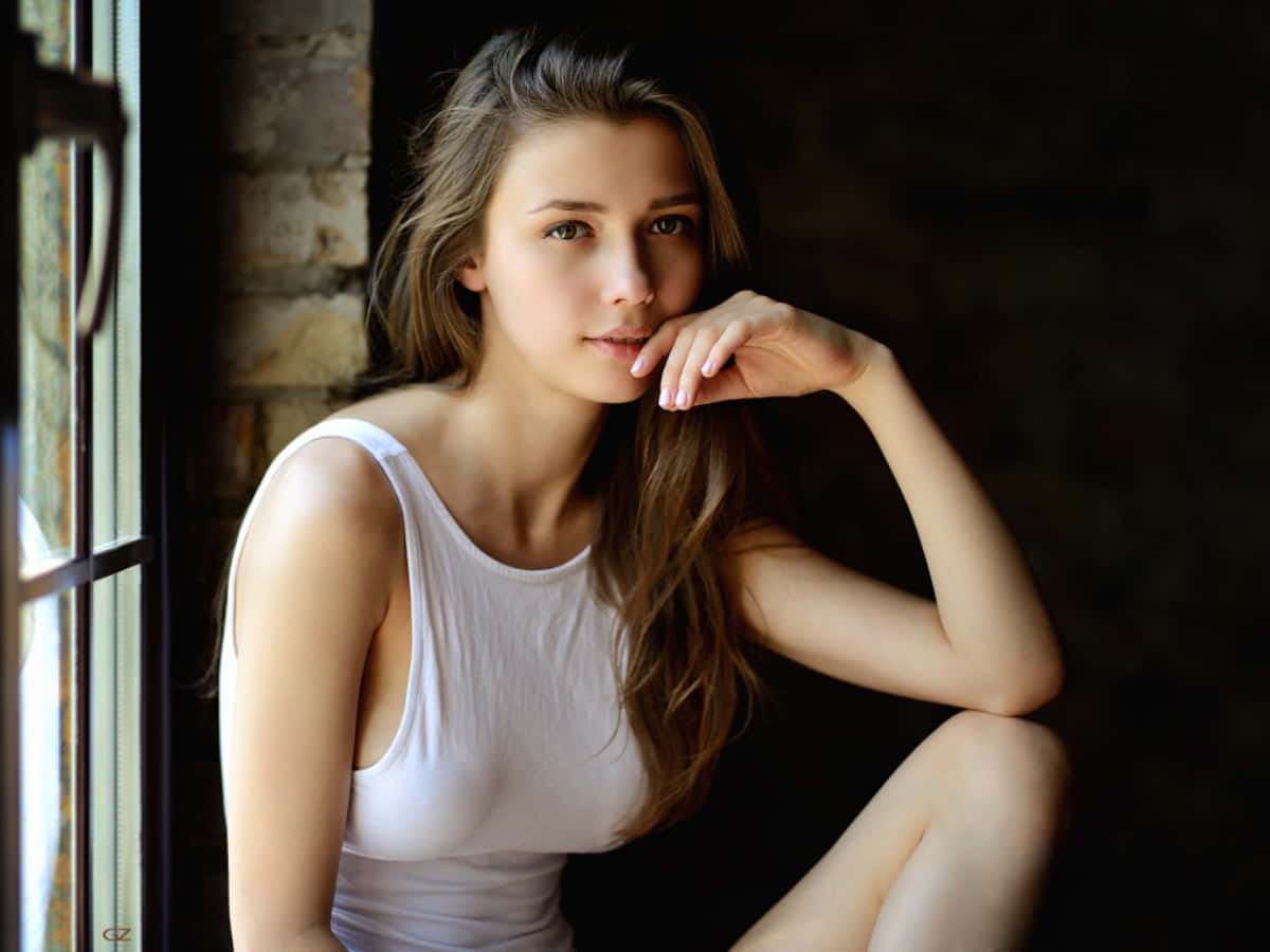 模特兒 桌布 壁紙 — 一位身穿白色背心的年輕女性坐在磚牆窗邊，她的手在柔和自然光下靠近嘴唇。