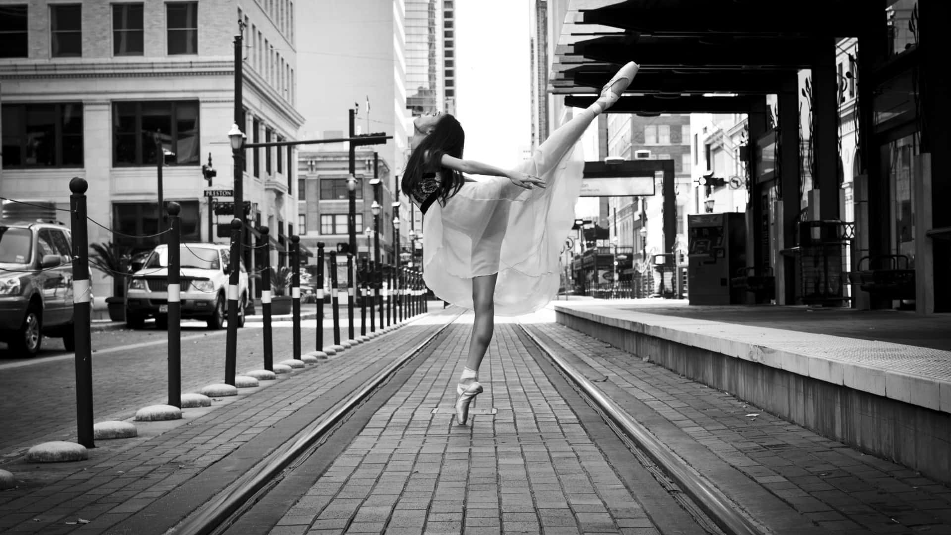 ballerina 桌布 壁紙 — 一位身穿白色舞衣的芭蕾舞者在城市街道的電車軌道上優雅地做出阿拉貝斯克舞姿，黑白照片。