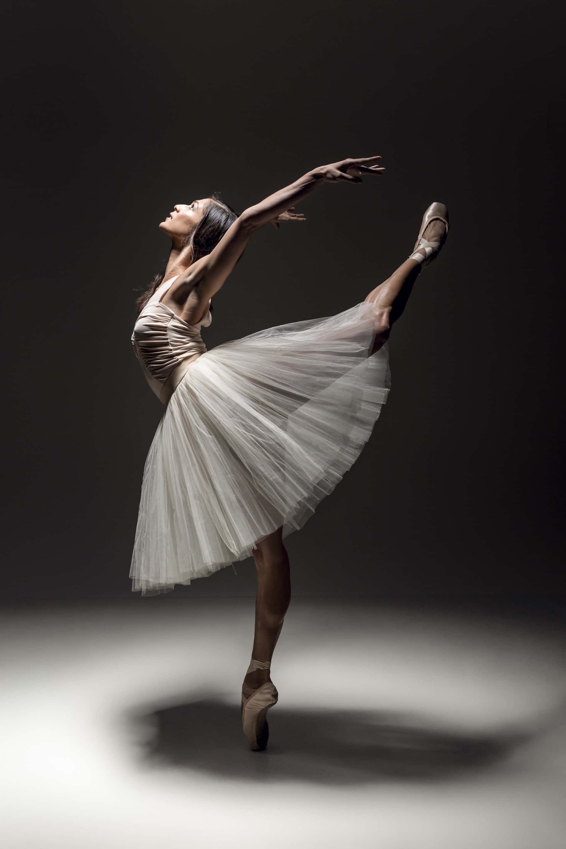 dancer 桌布 壁紙 — 一位身穿白色芭蕾舞裙的優雅芭蕾舞者，在昏暗的攝影棚背景下以足尖演繹優美的阿拉貝斯克姿勢。