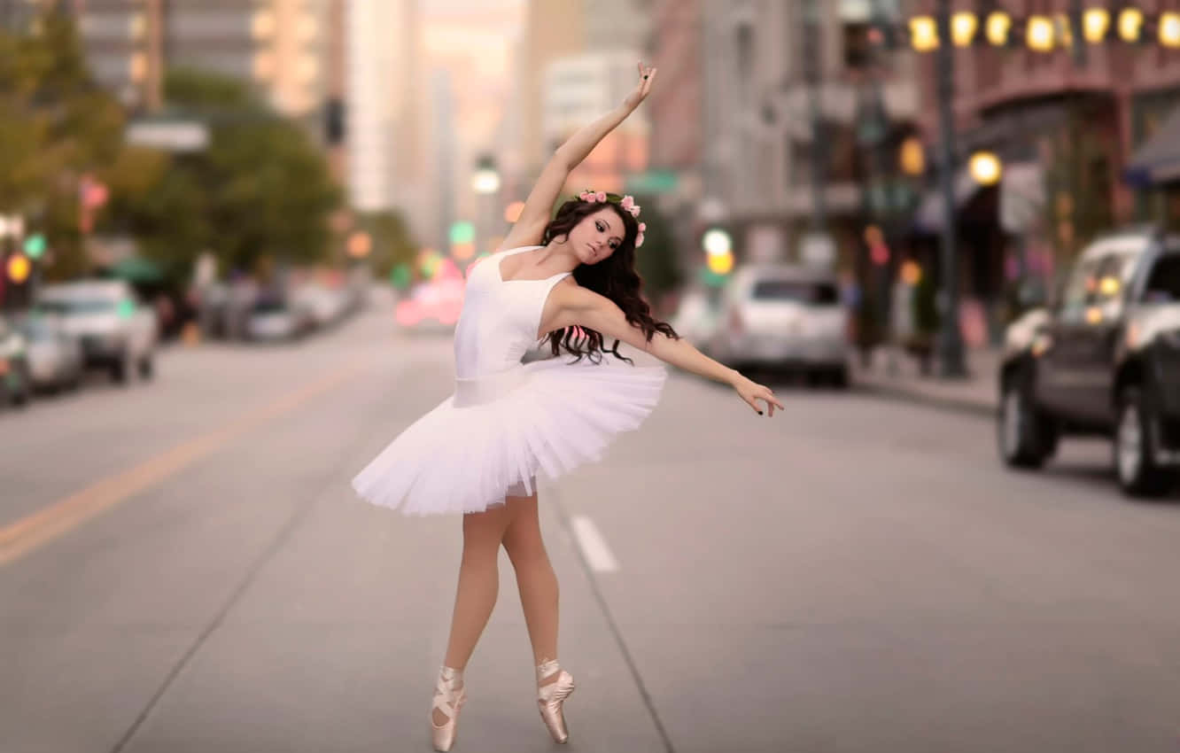 Ballet 桌布 壁紙 — 一位身穿白色芭蕾舞裙的芭蕾舞者在黃金時段的城市街道上優雅地踮起腳尖擺姿勢，身後是模糊的汽車和建築物。
