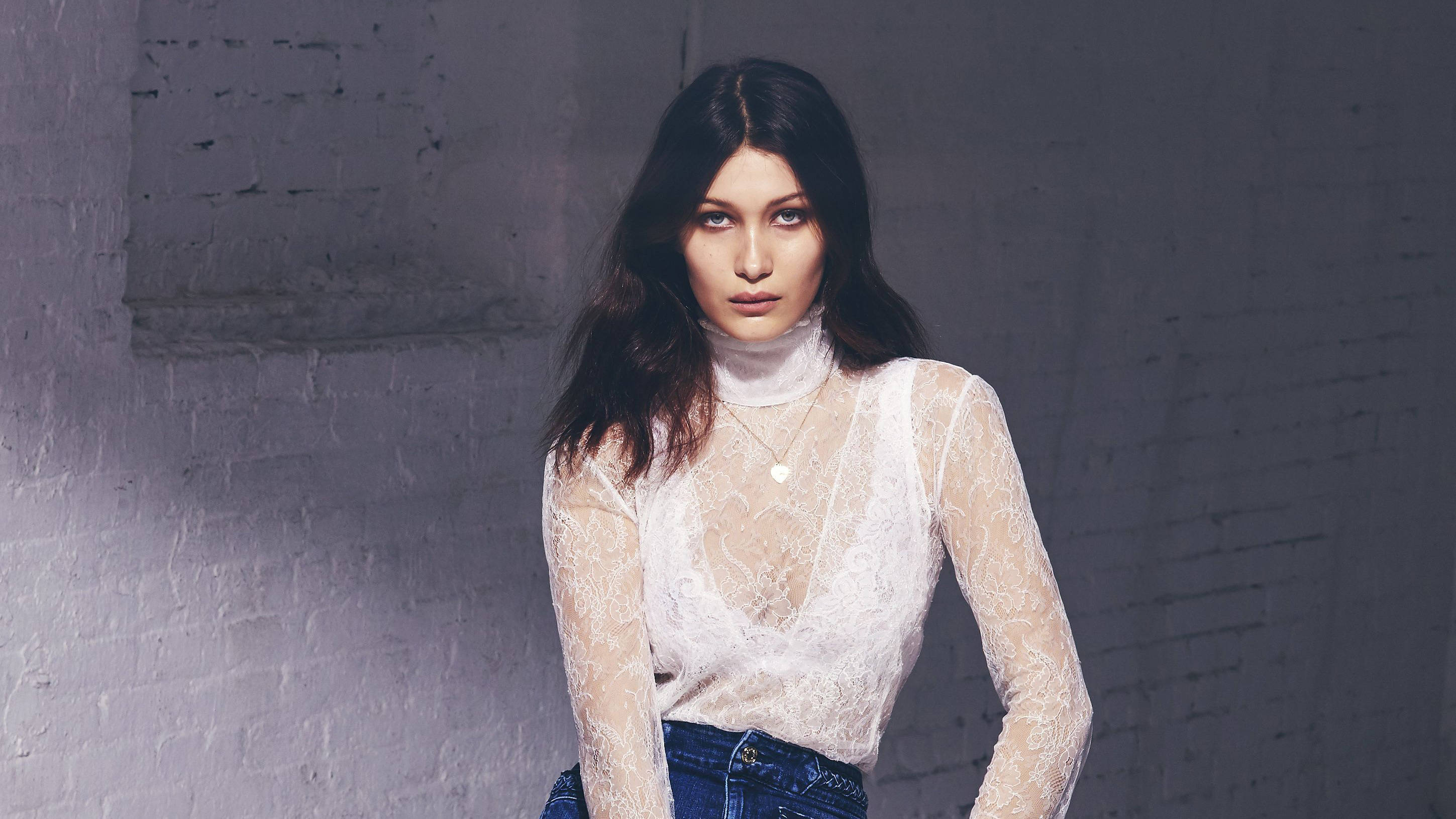 Bella Hadid 桌布 壁紙 — 一位身穿白色蕾絲上衣和藍色牛仔褲的女性，站在有紋理的白色磚牆前，光線柔和。