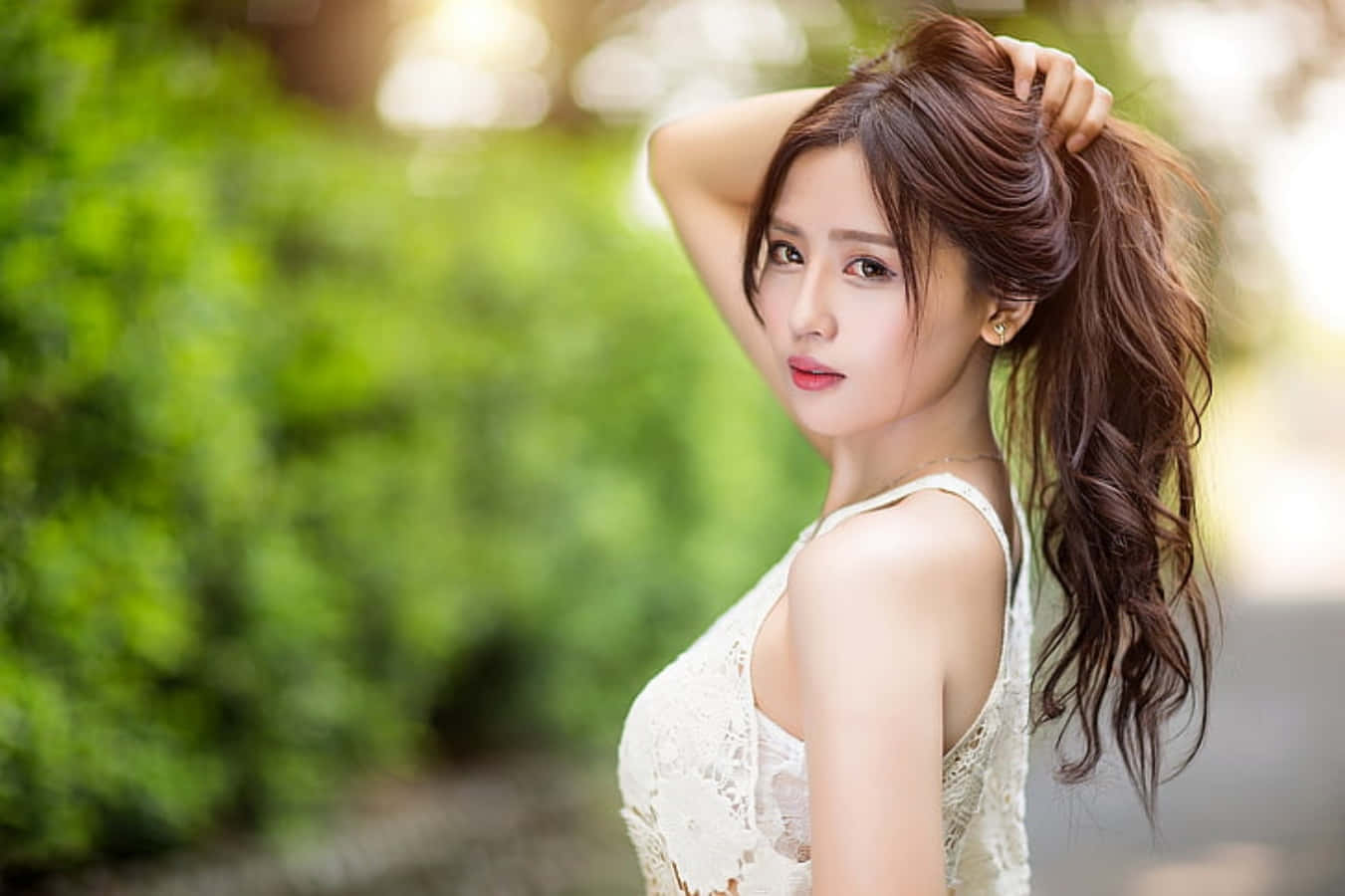 美女 桌布 壁紙 — 一位身穿白色蕾絲上衣的女性在戶外擺姿勢，一手放在頭髮上，背景是柔和的綠色模糊效果。