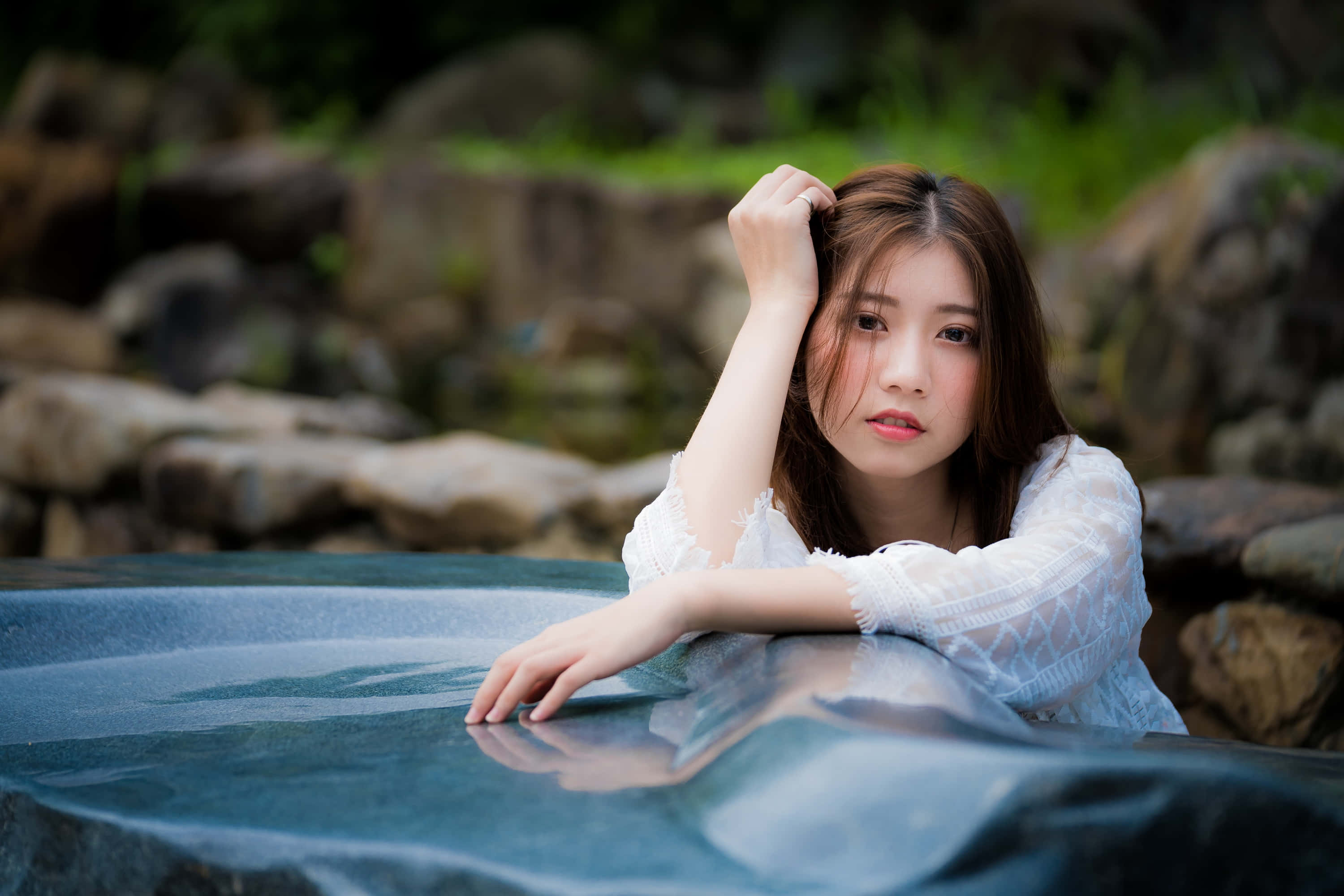 少女風 桌布 壁紙 — 一位身穿白色蕾絲上衣的年輕女子，將手臂擱在一個被苔蘚岩石和綠植環繞的反射水池上。