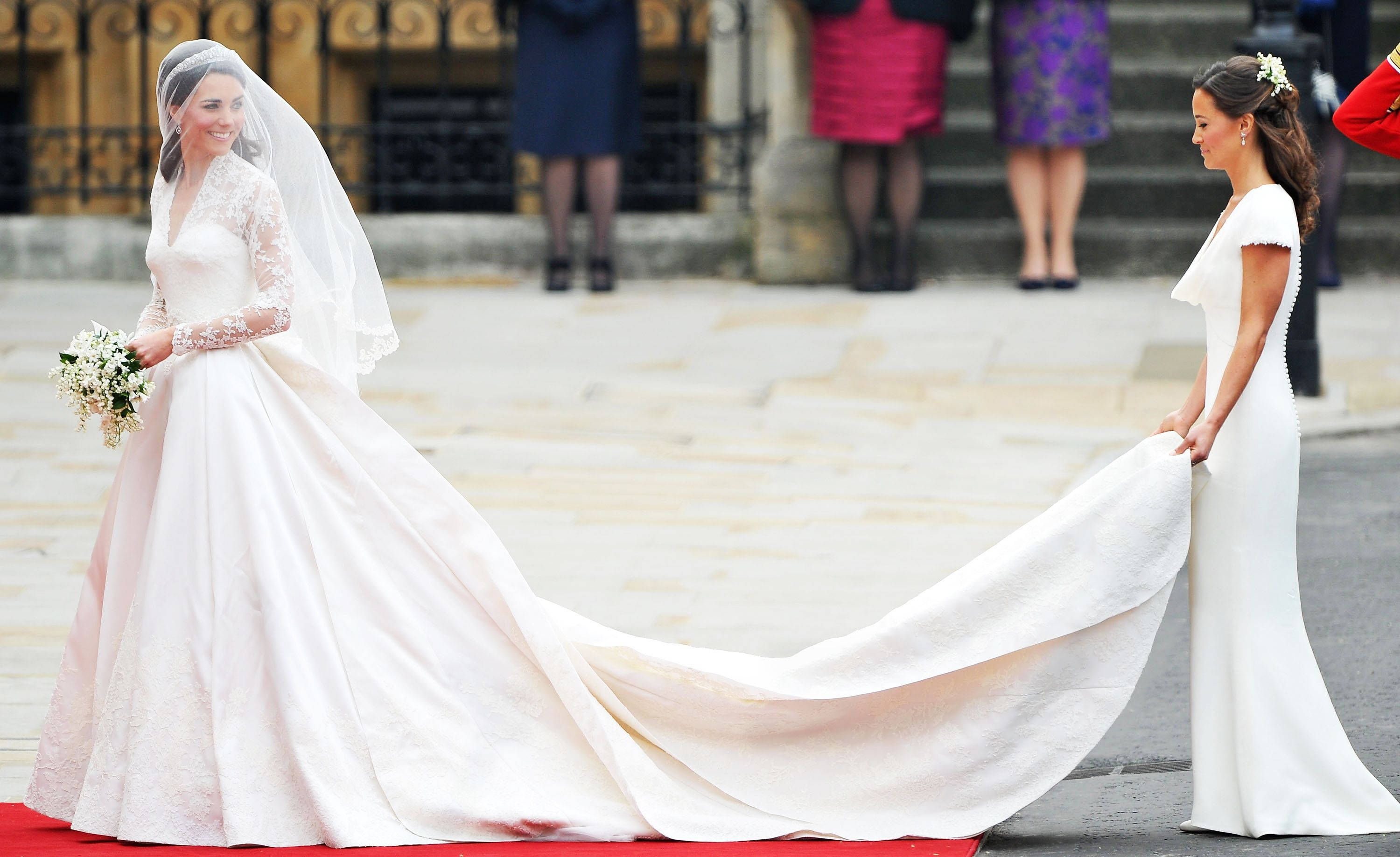 Royal 桌布 壁紙 — 一位身穿白色蕾絲婚紗、長長拖尾的新娘微笑著，一位身穿白色連衣裙的伴娘幫助她在一座宏偉建築外的紅地毯上行走。