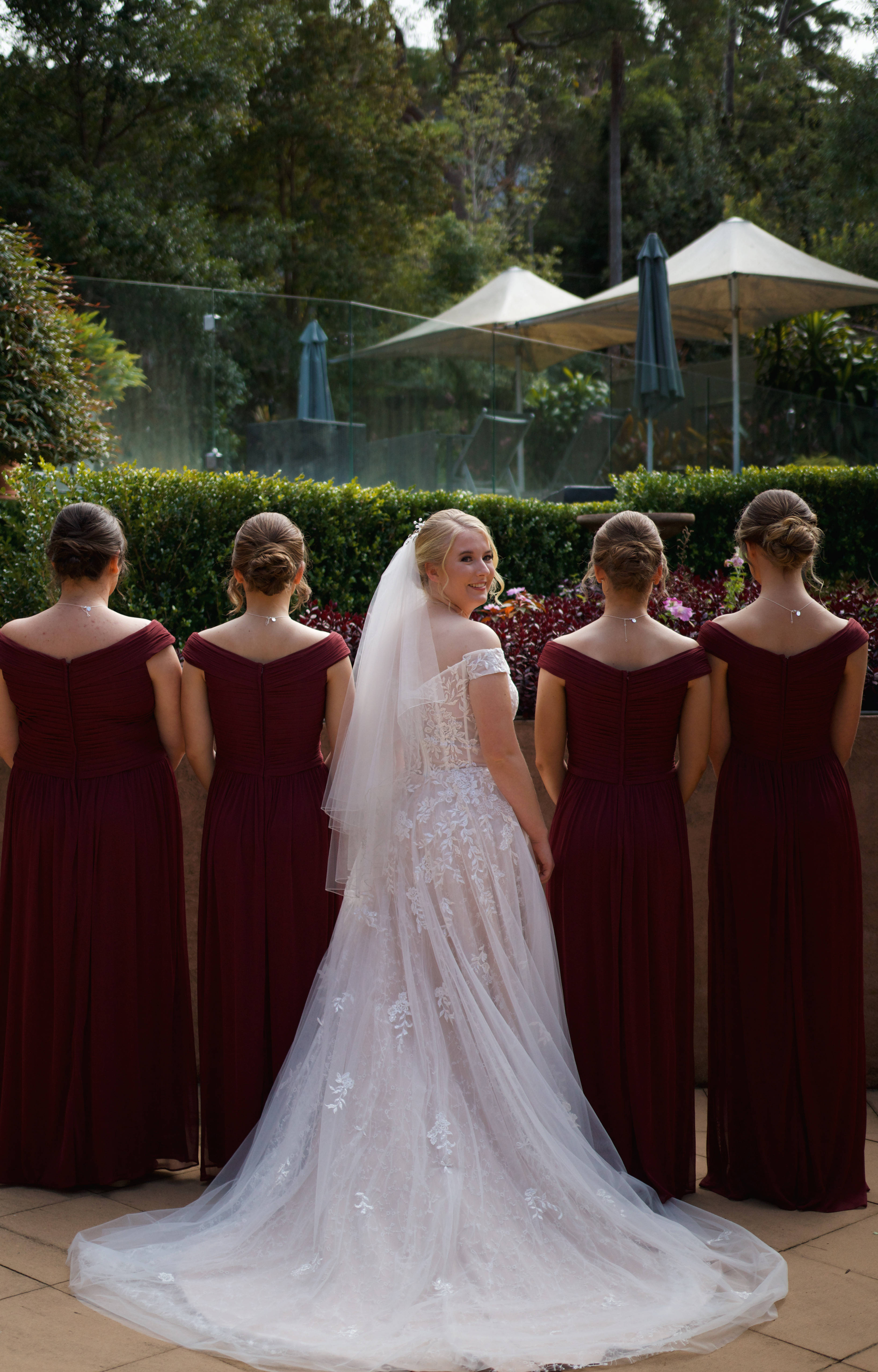 Bridesmaids 桌布 壁紙 — 一位身穿白色蕾絲婚紗、頭戴頭紗的新娘轉身微笑，四位身穿酒紅色禮服的伴娘站在她身後的庭園中。