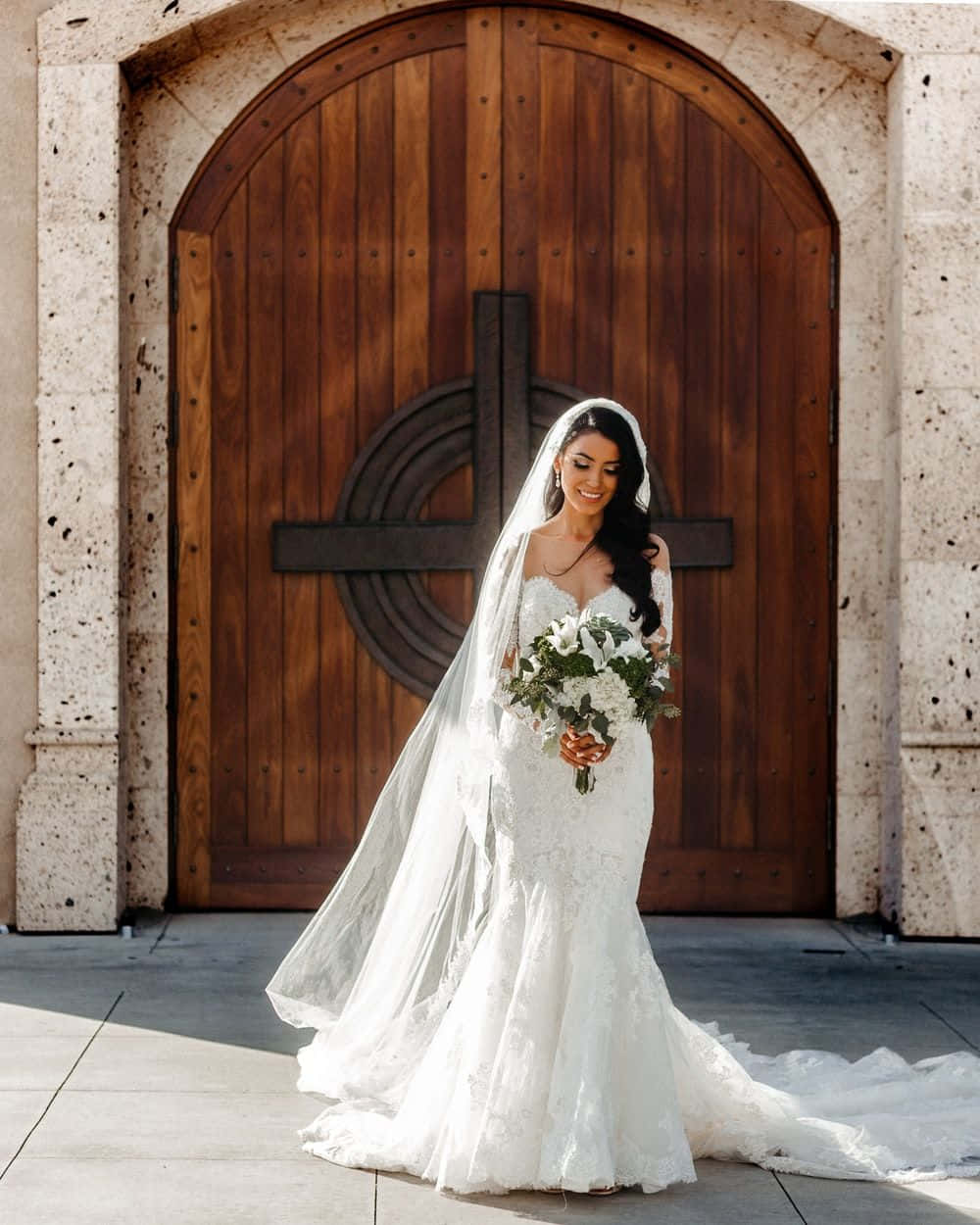 Bride 桌布 壁紙 — 一位身穿白色蕾絲婚紗和頭紗的新娘，微笑著站在帶有十字架的大型木製教堂門前。