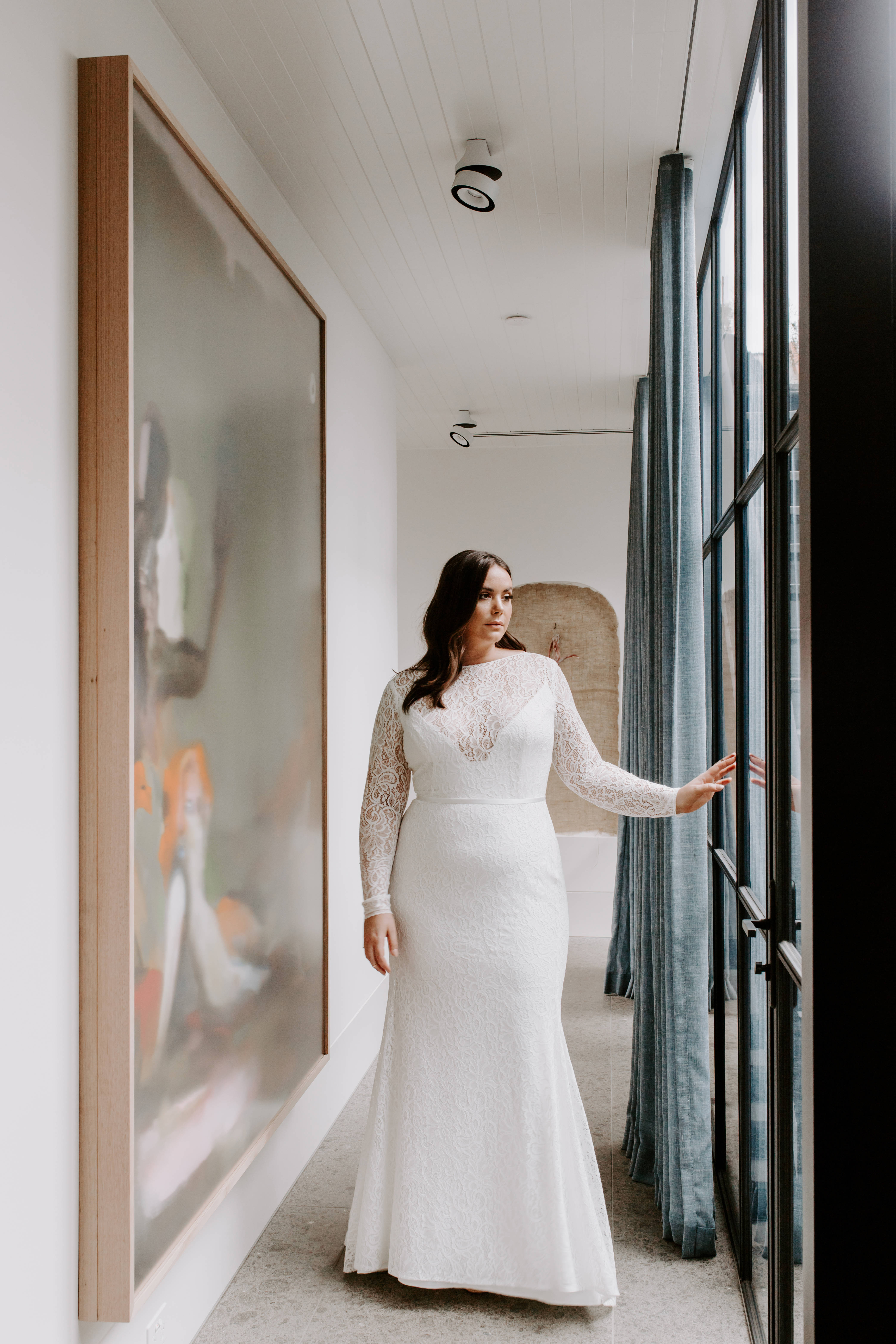 Wedding Aesthetic 桌布 壁紙 — 一位身穿白色蕾絲婚紗的女性站在現代走廊中，觸摸著大型抽象畫旁邊的玻璃門。