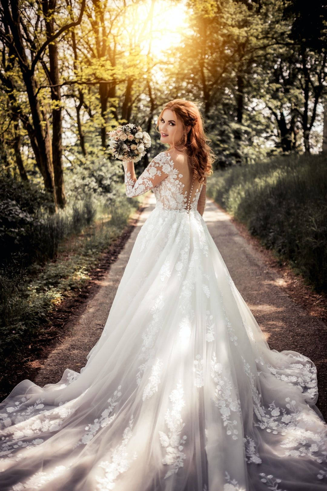 Bride 桌布 壁紙 — 一位身穿白色蕾絲婚紗的新娘，手持花束，走在陽光明媚的森林小徑上。