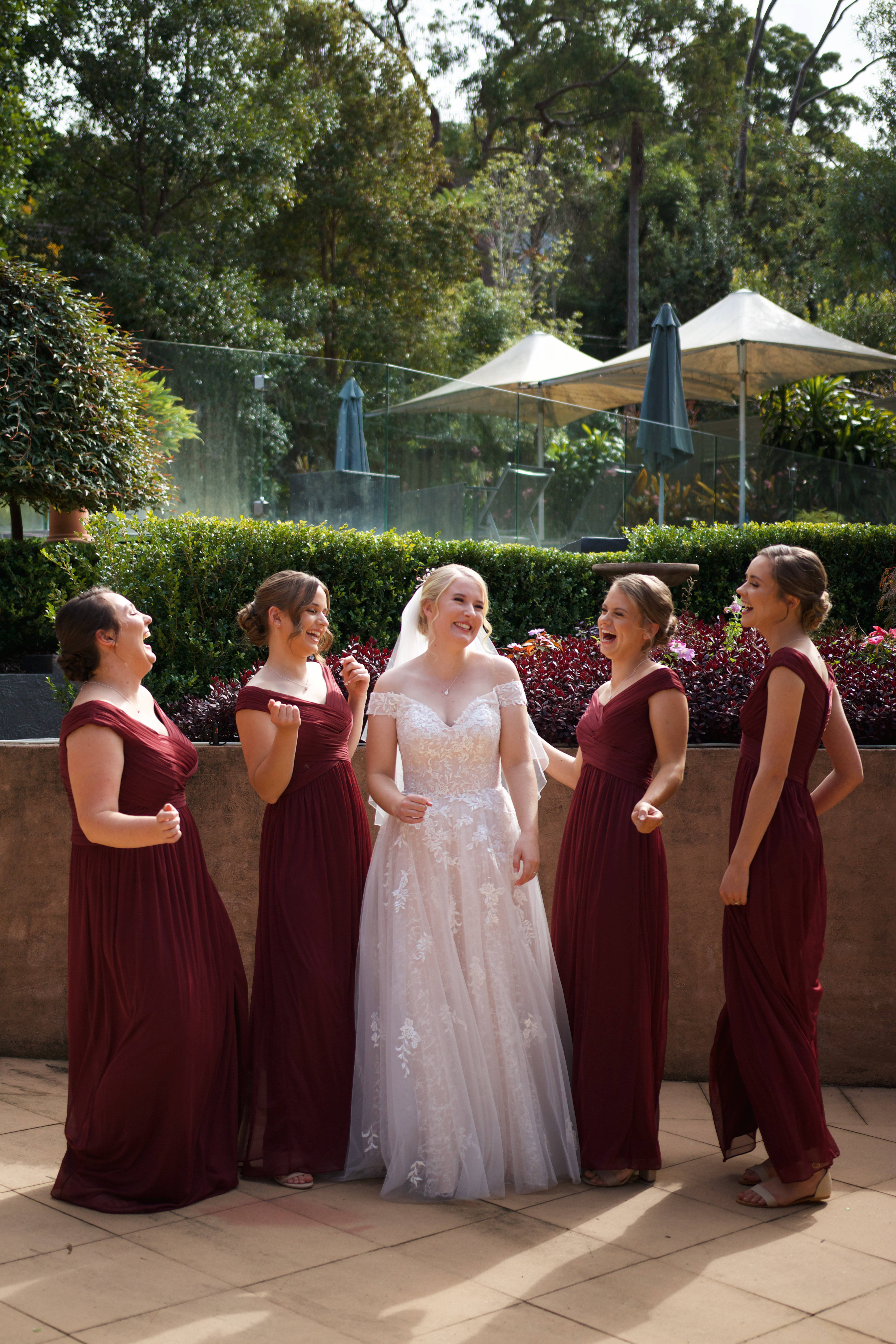 Bridesmaids 桌布 壁紙 — 一位身穿白色蕾絲婚紗的新娘與身穿酒紅色禮服的伴娘們在陽光下的戶外傘下歡笑。