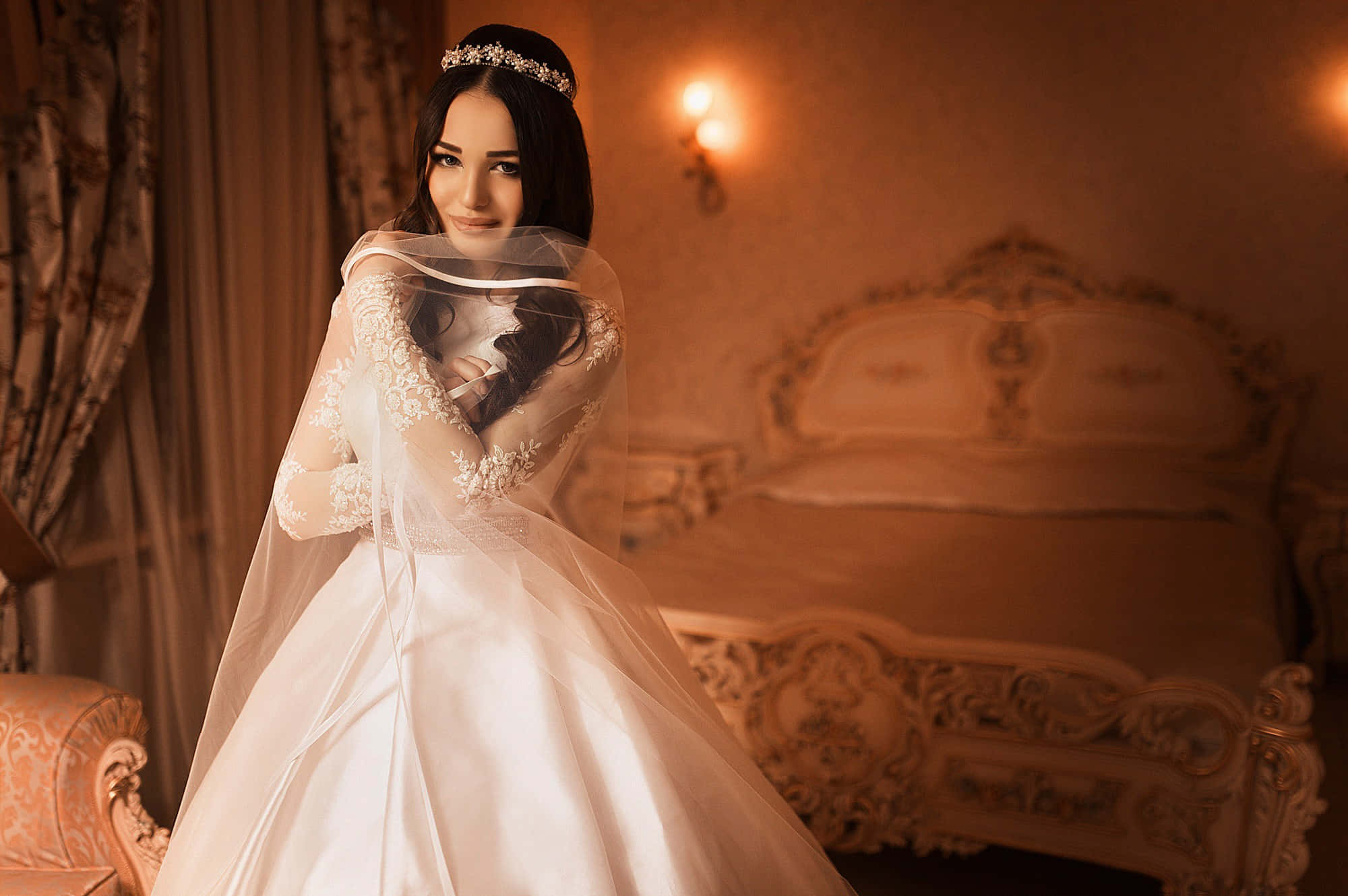 Bride 桌布 壁紙 — 一位身穿白色蕾絲禮服和頭紗的新娘，站在一個溫暖燈光、裝飾華麗的臥室裡，房間內有雕刻床和窗簾。