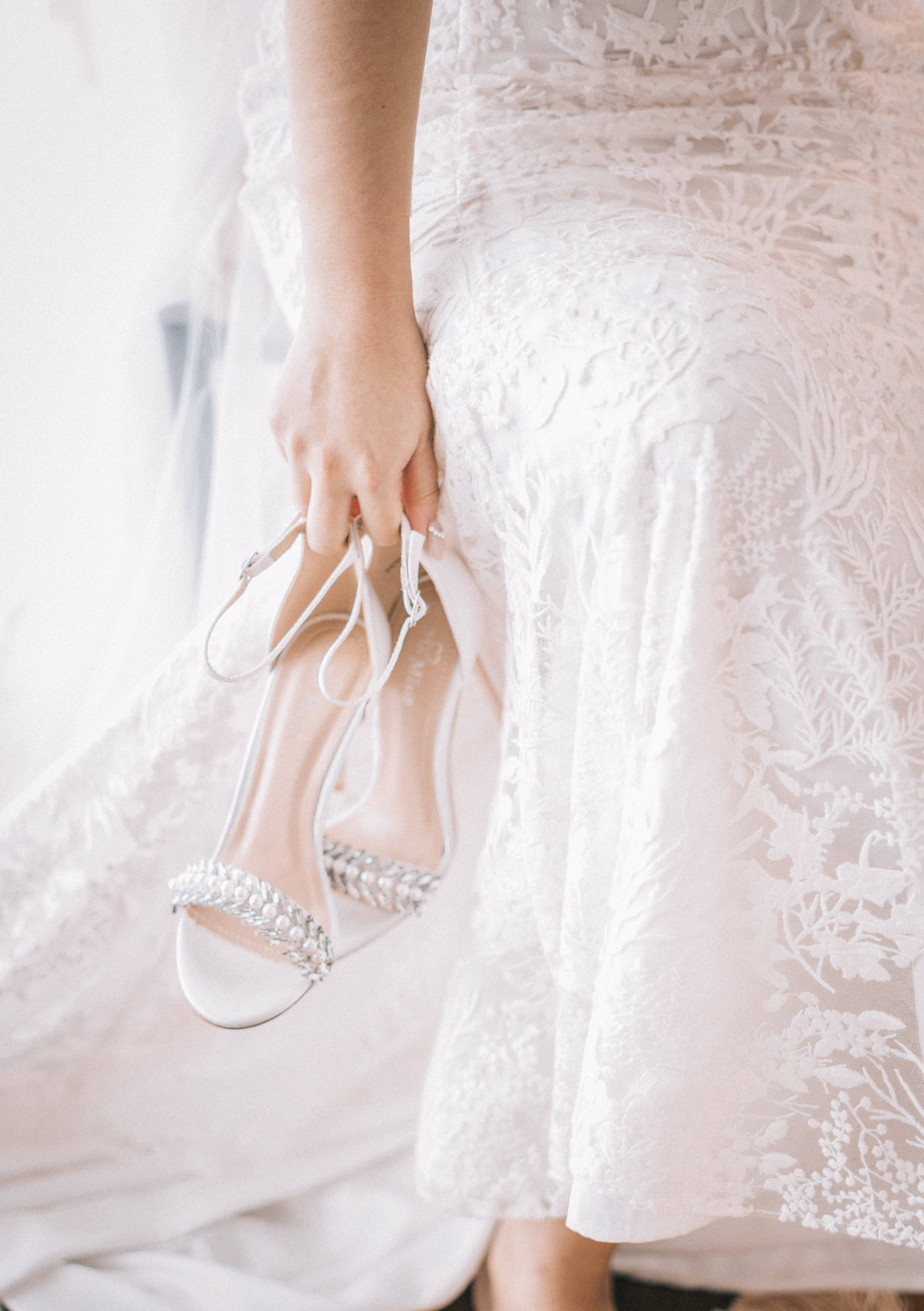Bridal 桌布 壁紙 — 一位身穿白色蕾絲禮服的新娘，在柔和自然光下拿著閃閃發光的銀色高跟鞋。