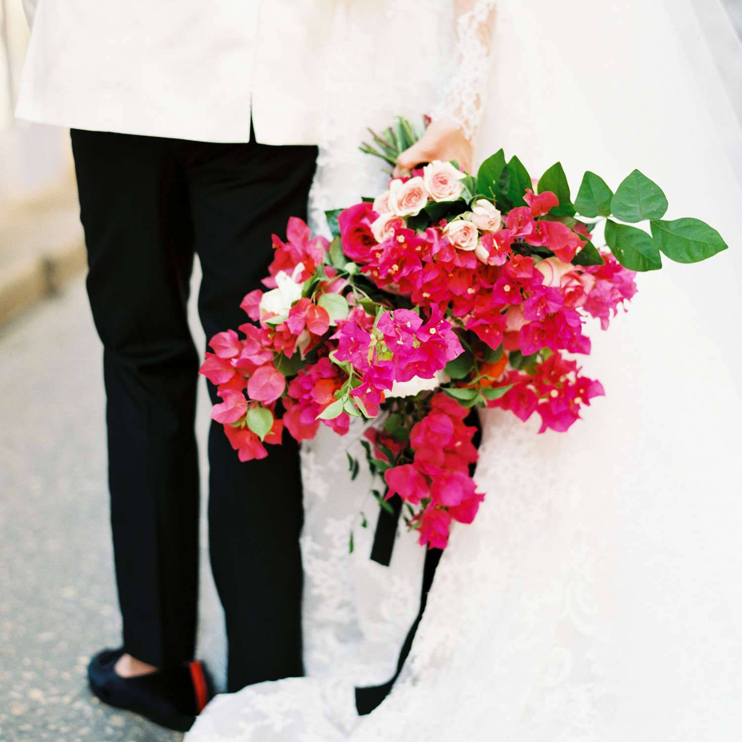 Wedding 桌布 壁紙 — 一位身穿白色蕾絲禮服的新娘，手持一束由粉紅色九重葛和淡粉玫瑰組成的鮮豔花束，旁邊是一位穿著黑色褲子和樂福鞋的新郎。
