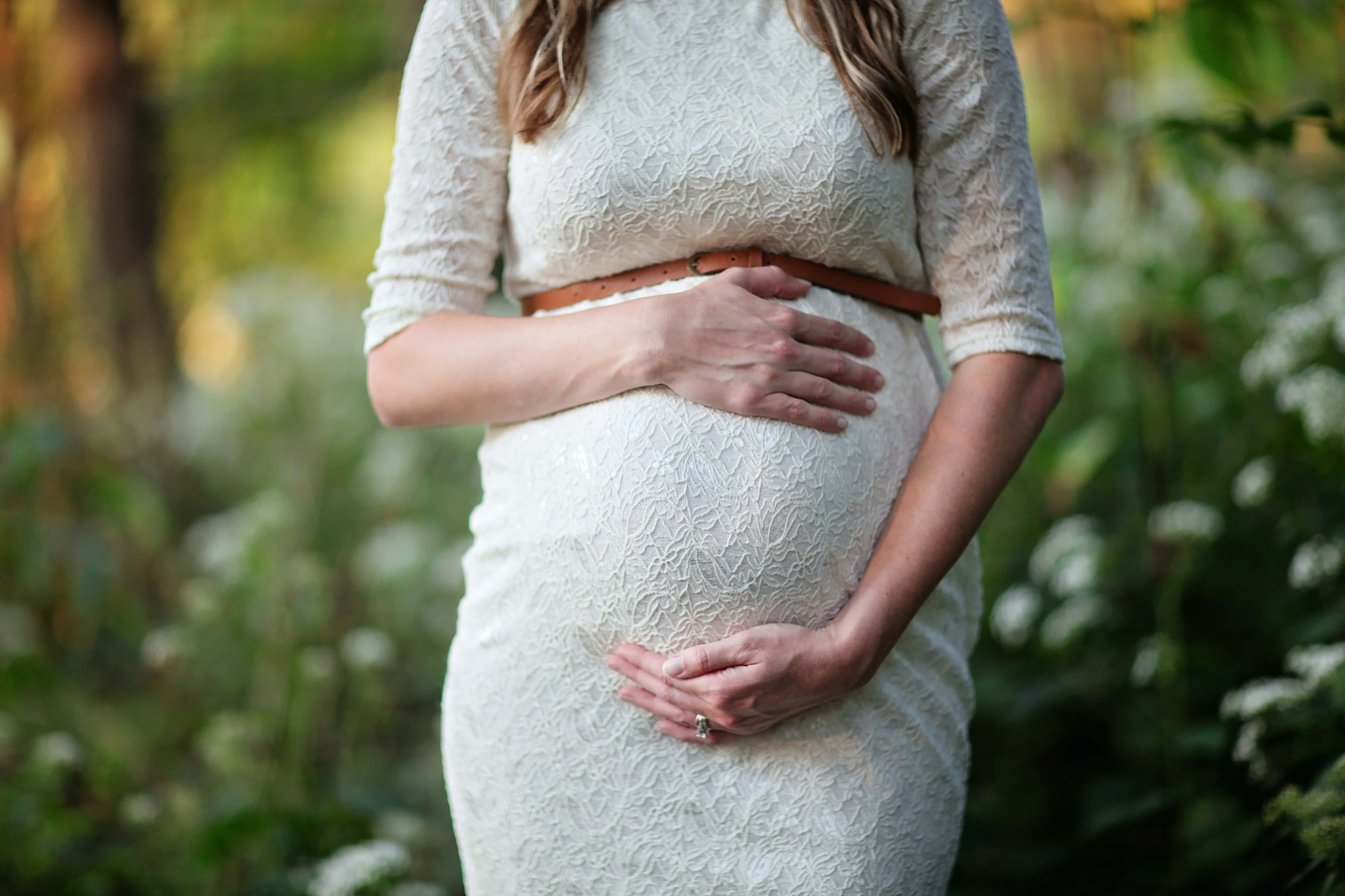 Pregnant 桌布 壁紙 — 一位身穿白色蕾絲裙的孕婦在柔軟的綠葉中，用雙手輕輕地捧著肚子。
