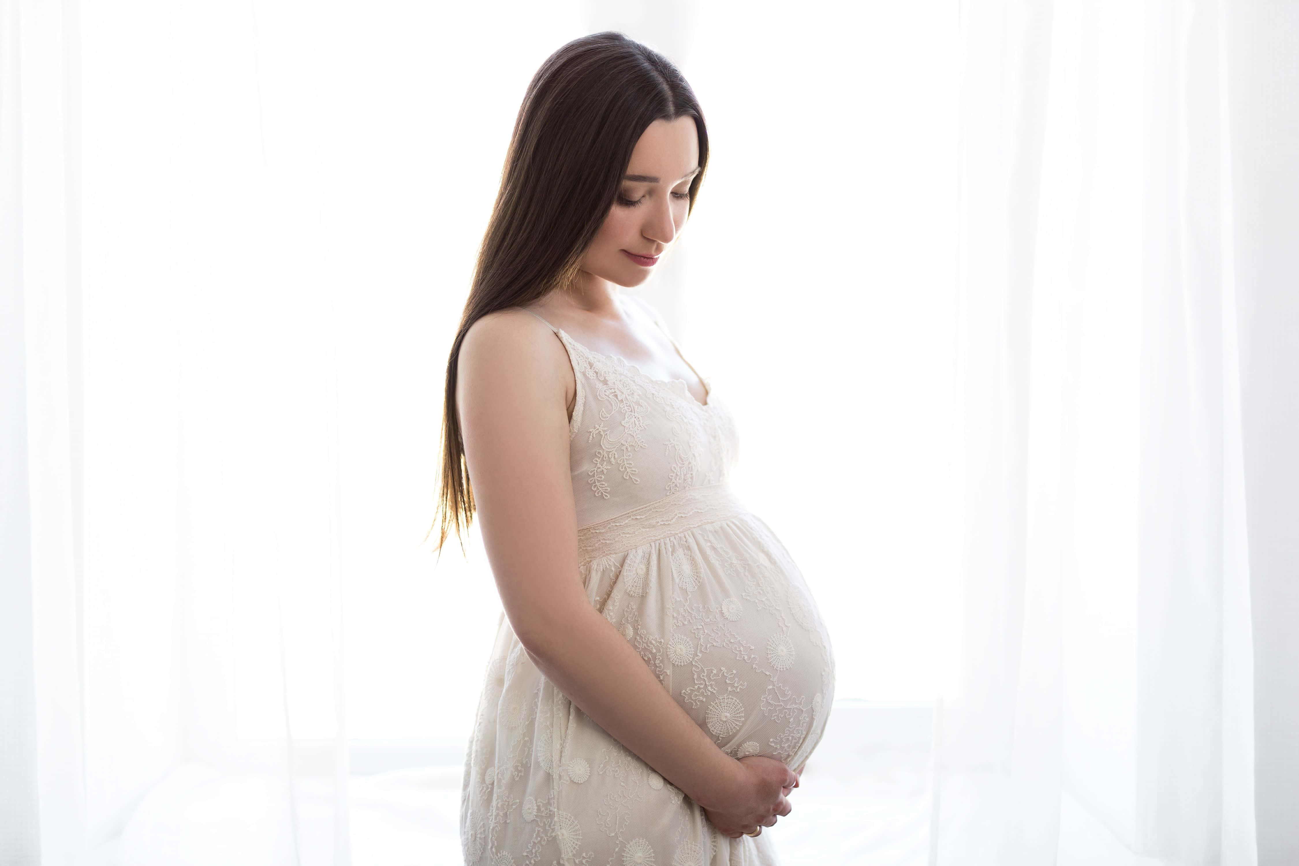 Pregnant 桌布 壁紙 — 一位身穿白色蕾絲裙的寧靜孕婦，在明亮的窗邊輕柔地抱著肚子，窗簾薄透。