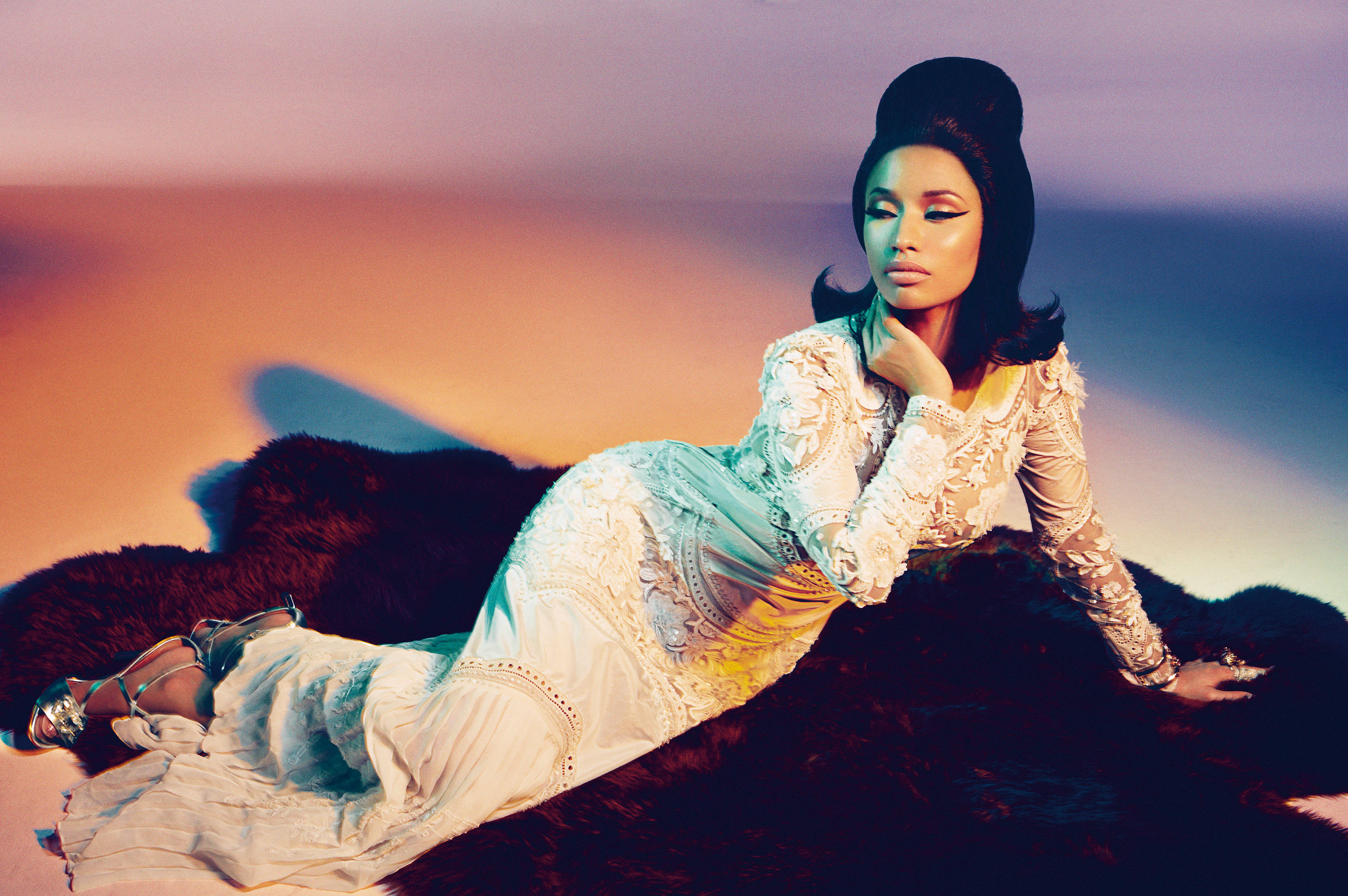 Nicki 桌布 壁紙 — 一位身穿白色蕾絲長袍的女性，在漸層日落天空的背景下，斜倚在深色毛皮地毯上。